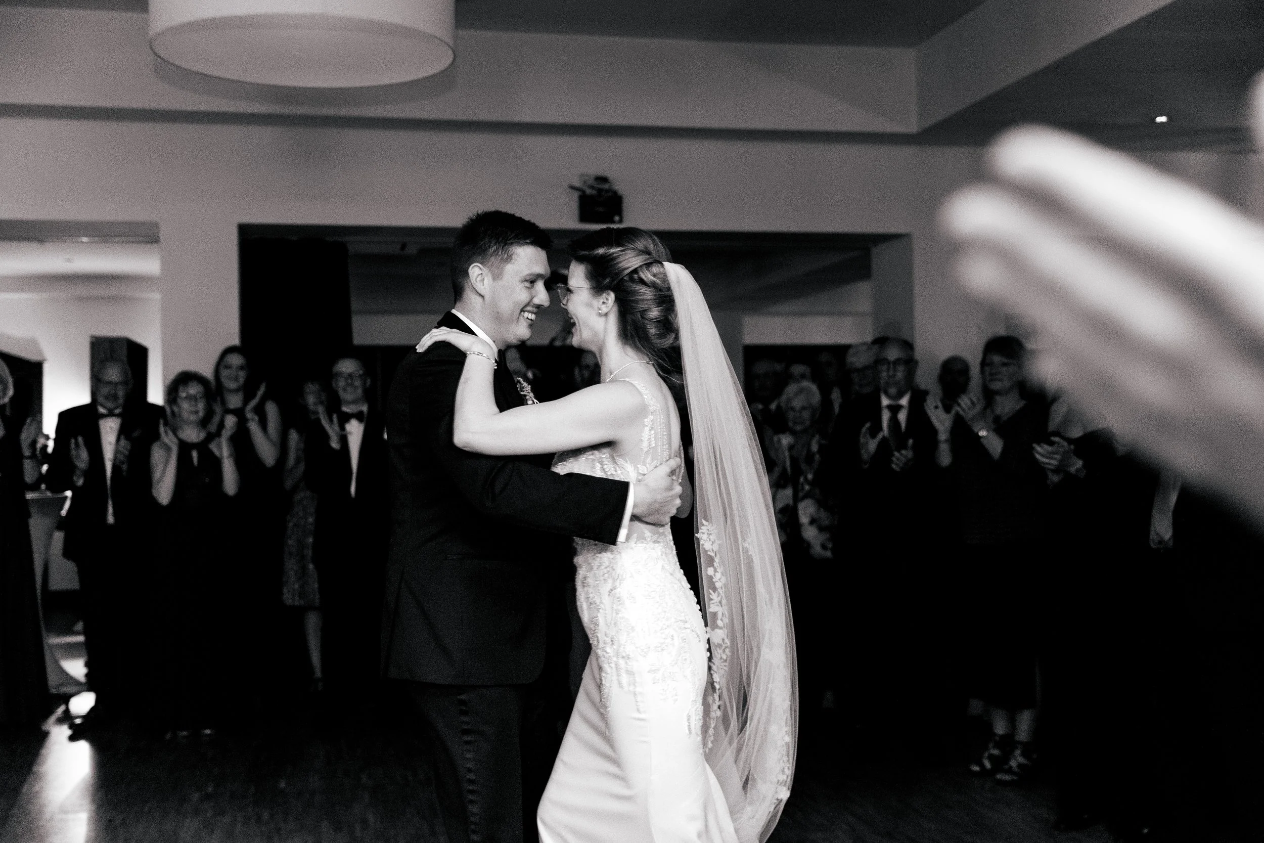 Braut und Bräutigam tanzen bei ihrer Hochzeit, umgeben von Gästen, in schwarz-weiß aufgenommen.
