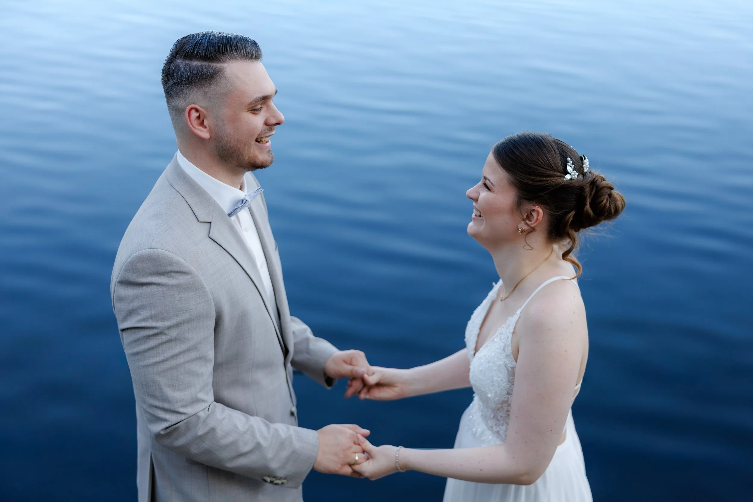 Ein Hochzeitspaar hält sich an einem Gewässer die Hände und lächelt sich an.