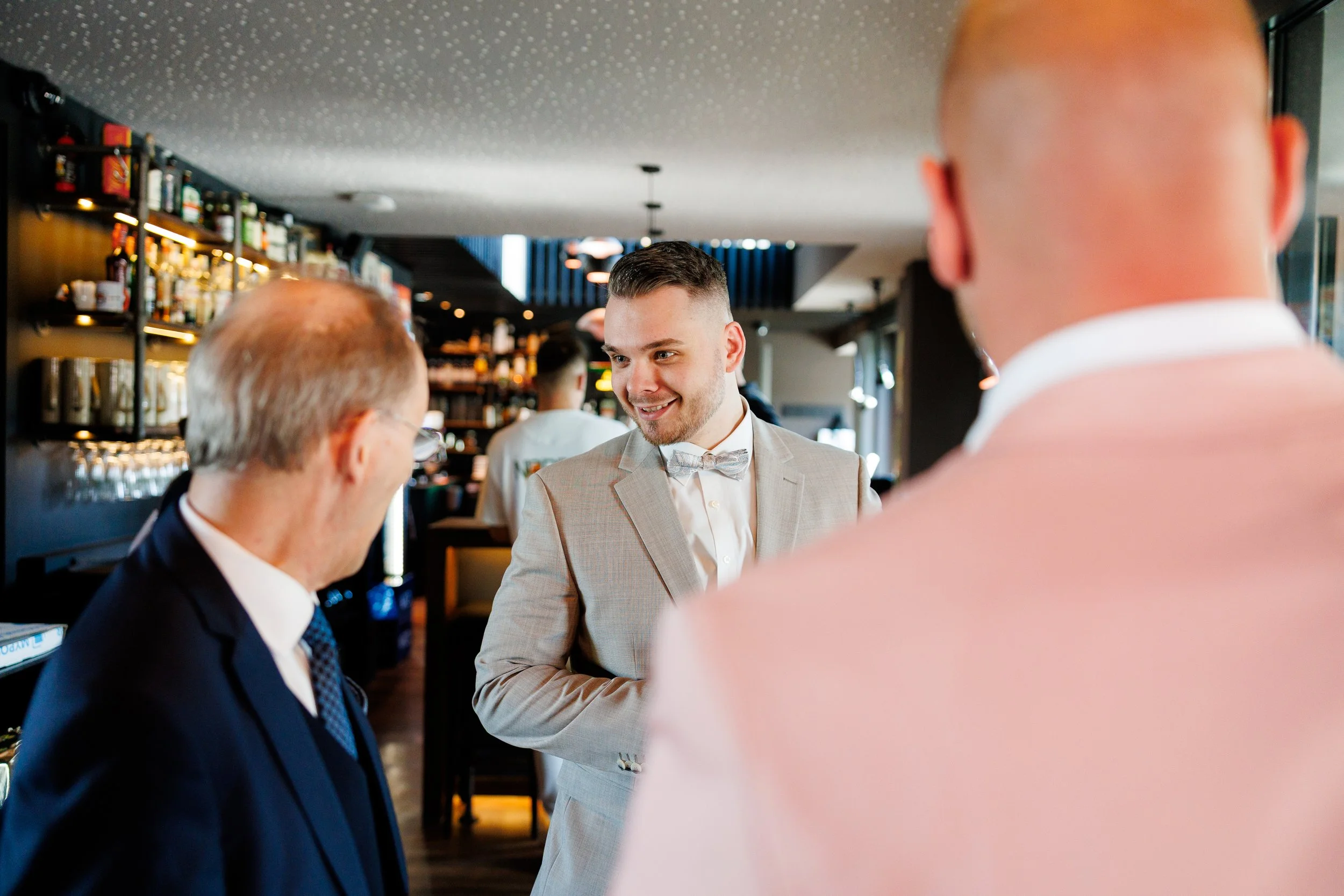 Mehrere Männer in einem Restaurant, unterhalten sich, im Hintergrund eine Bar mit Flaschen und Gläsern, einer trägt einen grauen Anzug mit Fliege.