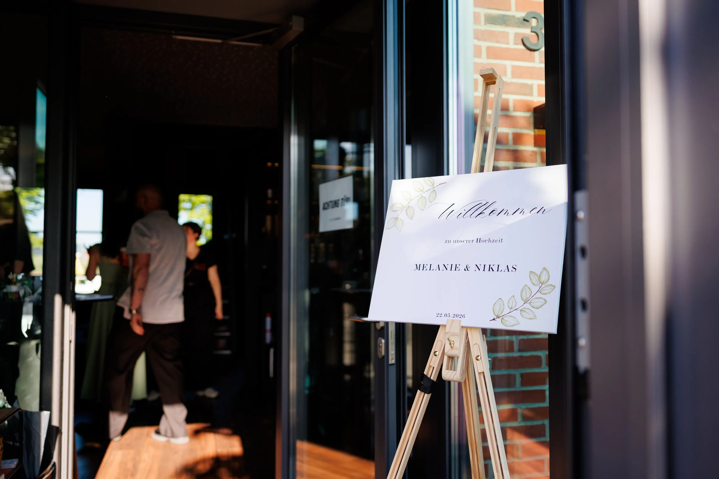 Ein Willkommensschild für eine Hochzeit mit den Namen Melanie und Niklas, Datum 22.05.2026, auf einer Staffelei vor einer Eingangstür bei einer Feier.