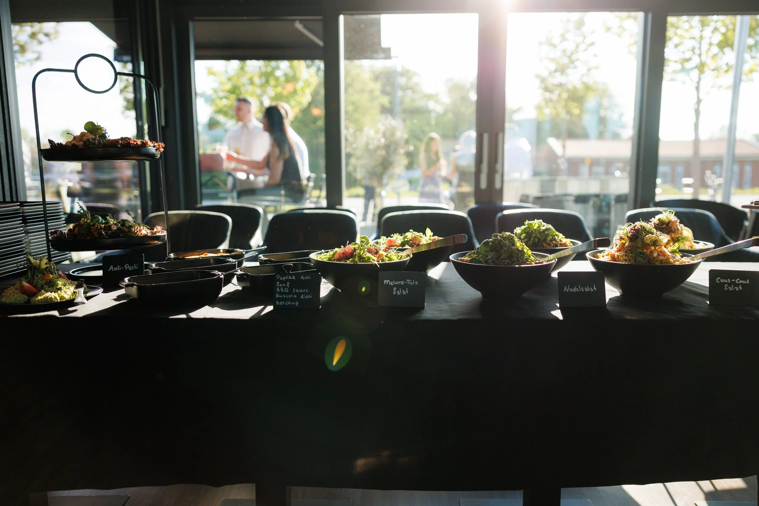 Buffet mit verschiedenen Salaten in Schüsseln auf einem Tisch, im Hintergrund große Fenster mit Blick nach draußen, wo Menschen im Sonnenlicht stehen.