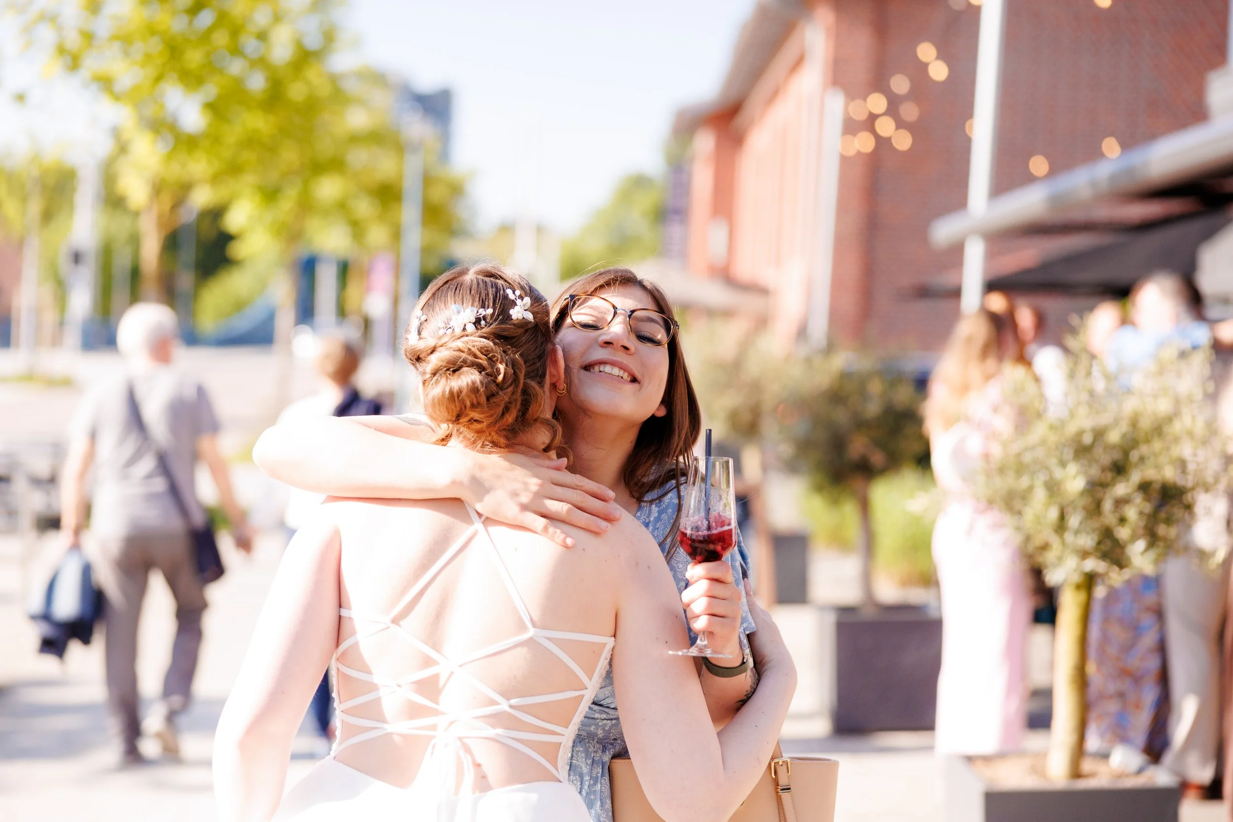 Zwei Frauen umarmen sich bei einer Feier im Freien, eine hält ein Glas mit rotem Getränk. Im Hintergrund sind weitere Menschen und bunte Bäume zu sehen.
