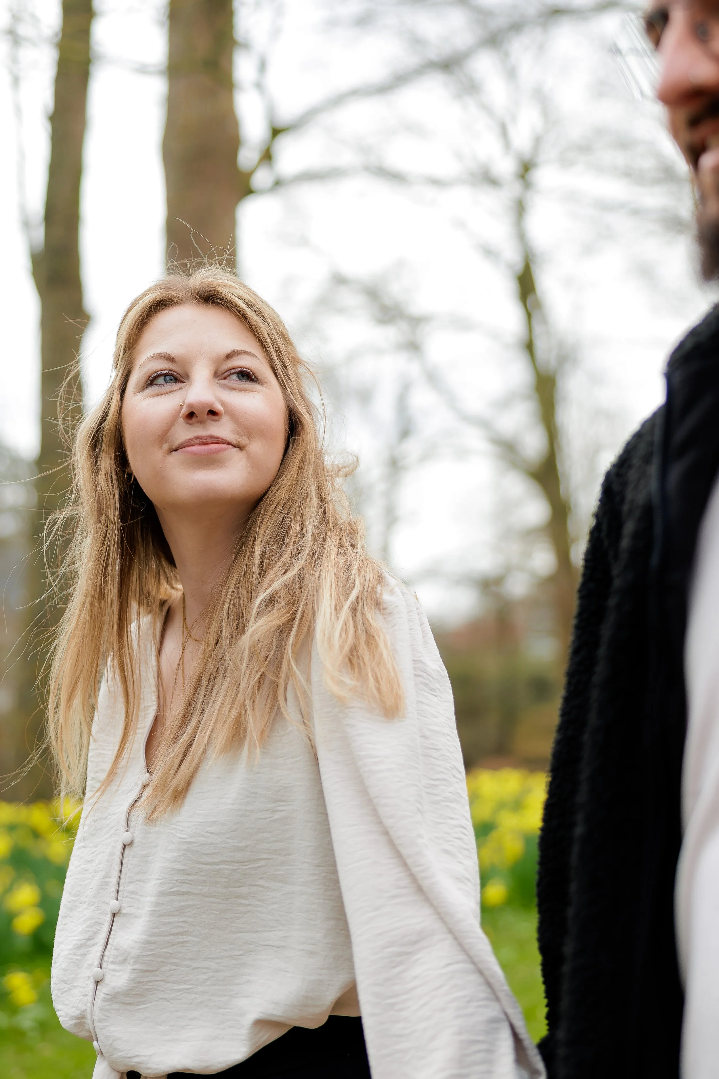 Eine junge Frau mit langen, lockigen blonden Haaren steht im Freien in einem Park, lächelnd und blickt zur Seite, während jemand anderes neben ihr steht, nur teilweise sichtbar.