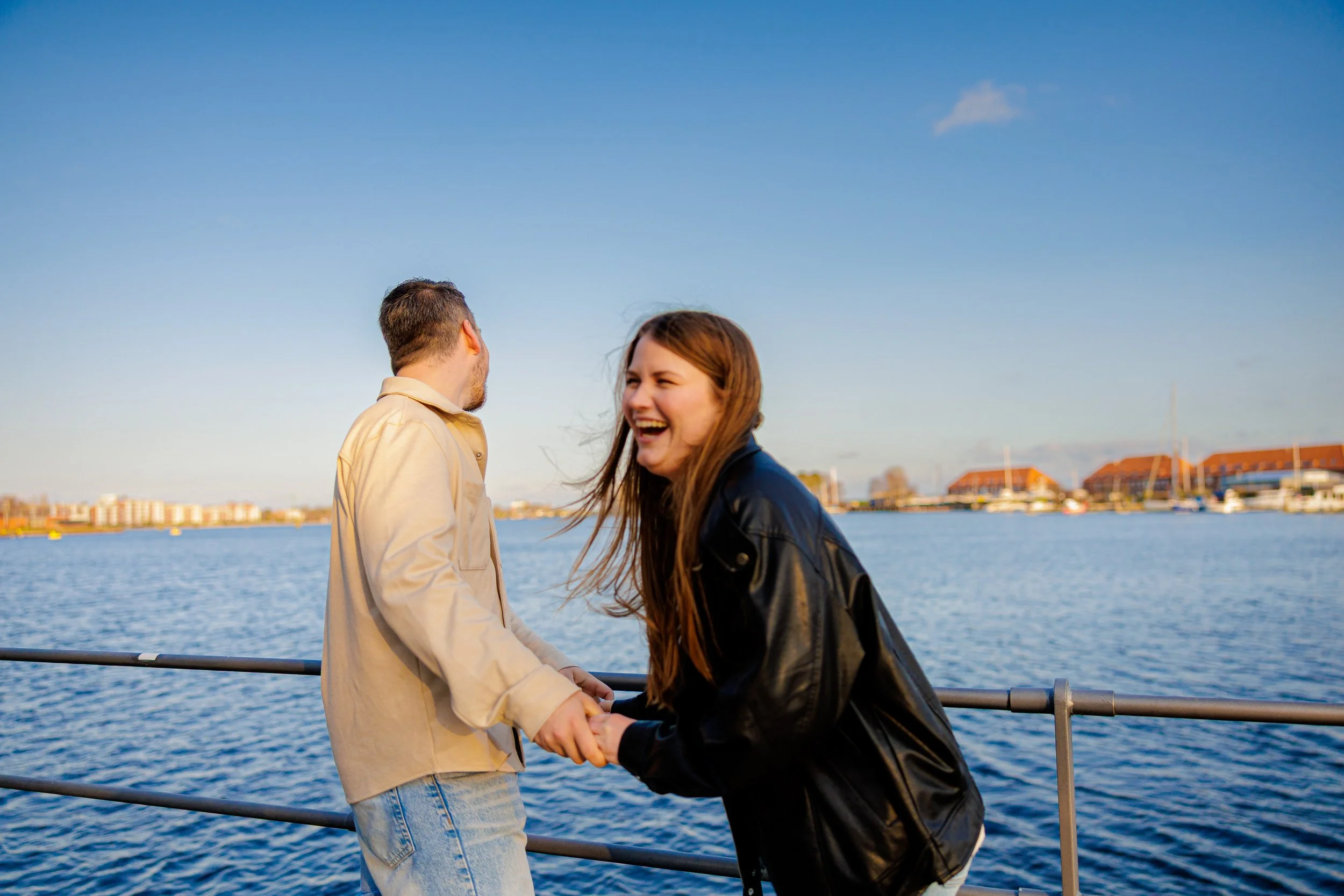 Ein lachendes Paar steht sich am Hafen bei Sonnenuntergang gegenüber, während sie Händchen halten und Blickkontakt haben, mit Wasser und Gebäuden im Hintergrund.