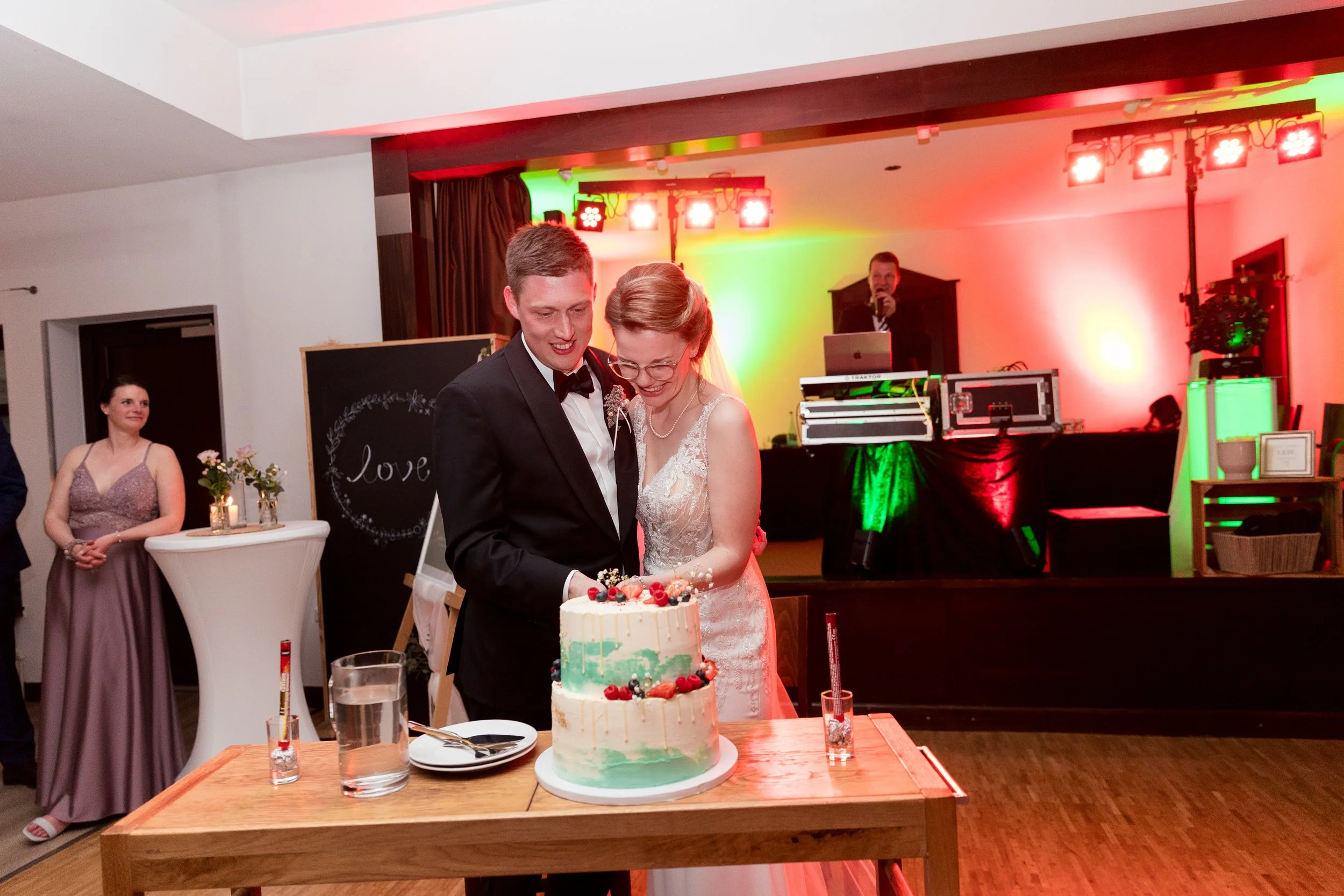 Ein Brautpaar schneidet eine Hochzeitstorte an, während im Hintergrund eine Person mit einem Mikrofon steht. Es gibt weitere Gäste und eine Tanzfläche mit bunter Beleuchtung.