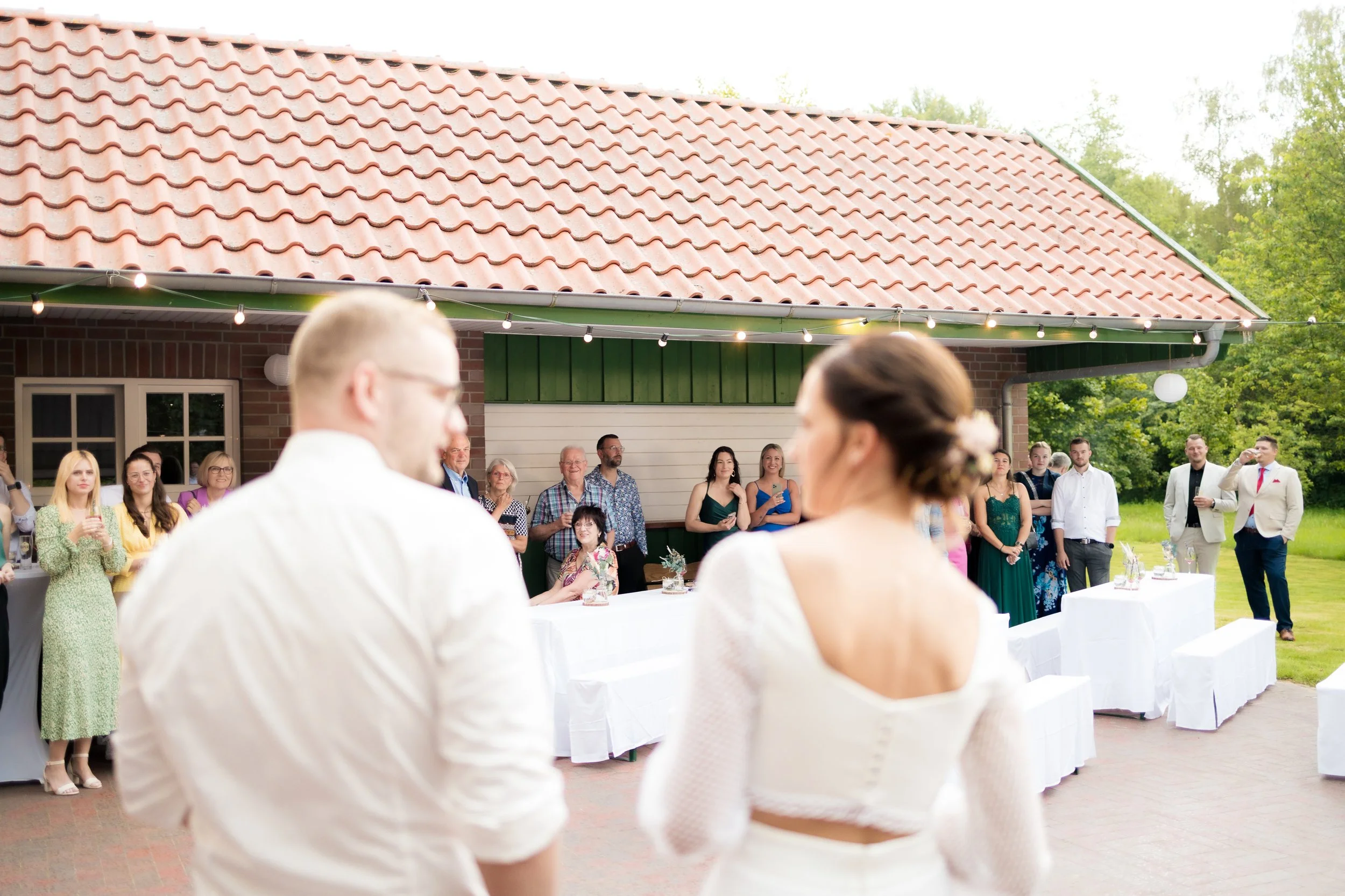 Viele Menschen bei einer Hochzeitsfeier im Freien, mit Paar im Vordergrund, bei einer Hochzeit am Tisch im Garten, im Sommer, mit Lampen und grüner Umgebung.