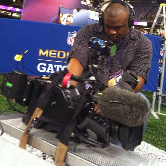 Four people standing on a rooftop with solar panels, discussing or examining the setup. The rooftop is covered with greenery, and a building with a pitched roof and chimney is visible in the background under a clear blue sky.