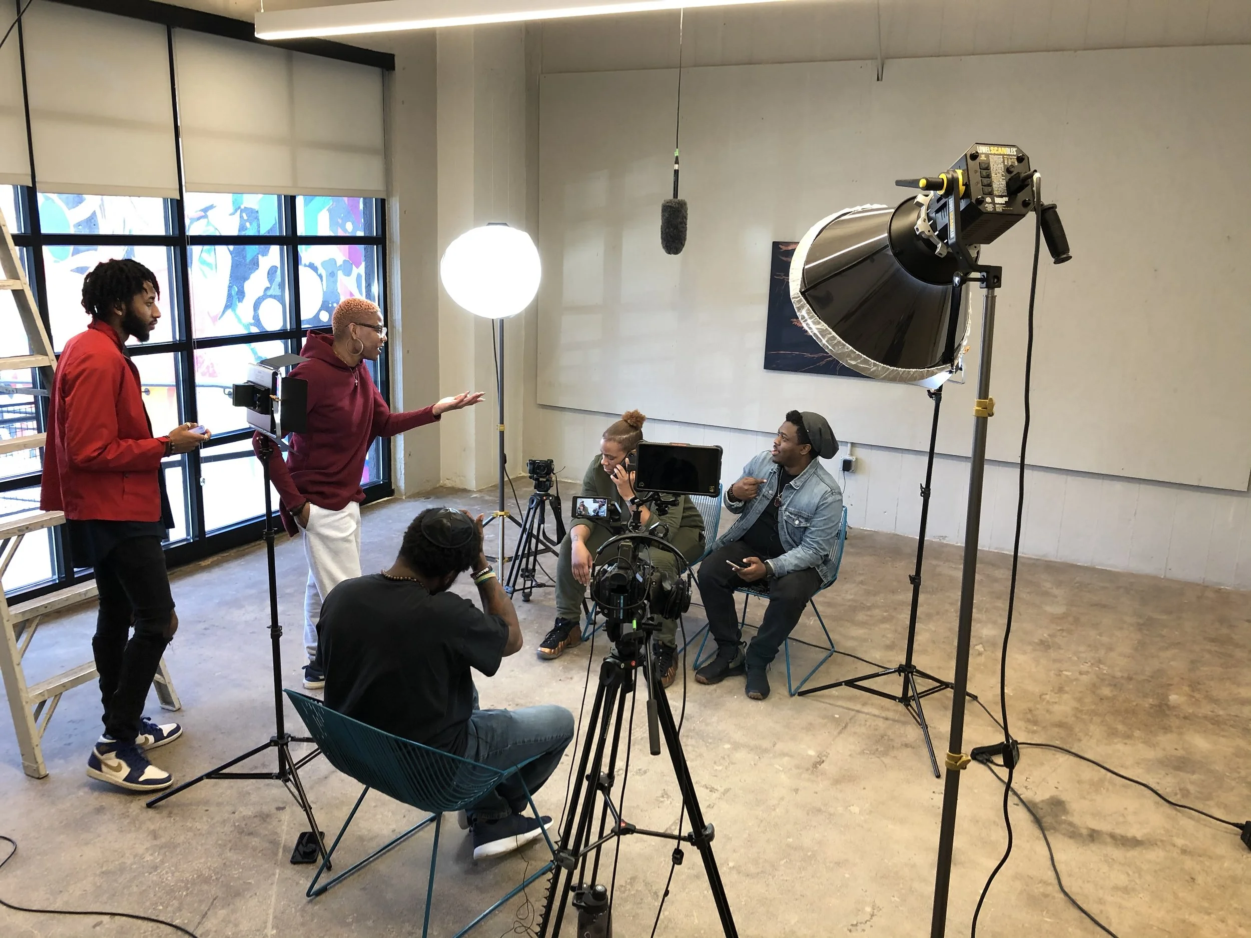 A group of seven people, six women and one man, sitting on stools in an office environment, with large windows in the background. They are dressed in business and casual attire. "Dream Big" and "Don't Give Up" posters are visible on the walls.