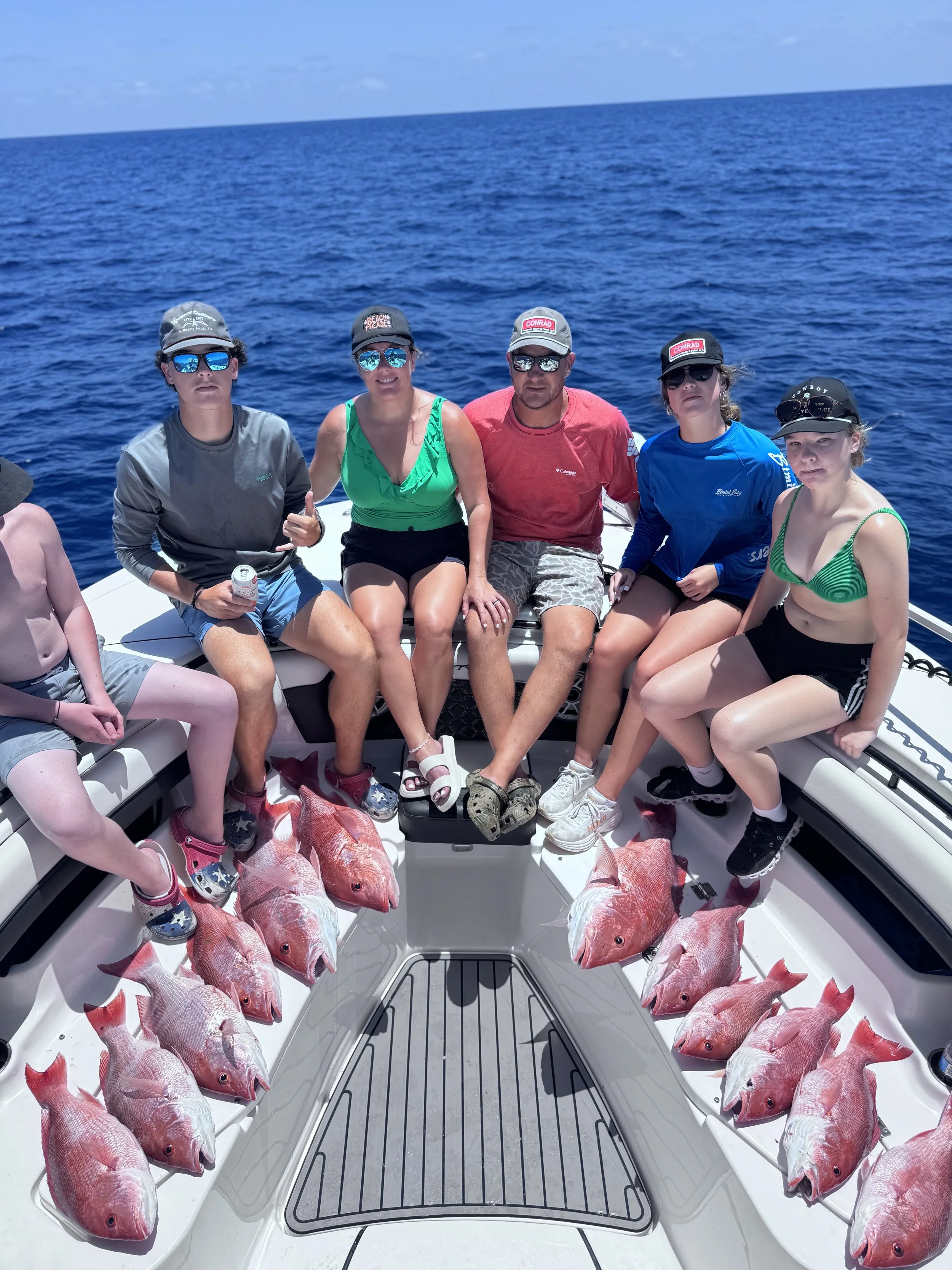 Three men on a boat holding a large fish they caught, with the ocean and sky in the background. They are smiling and wearing sunglasses and casual clothing.