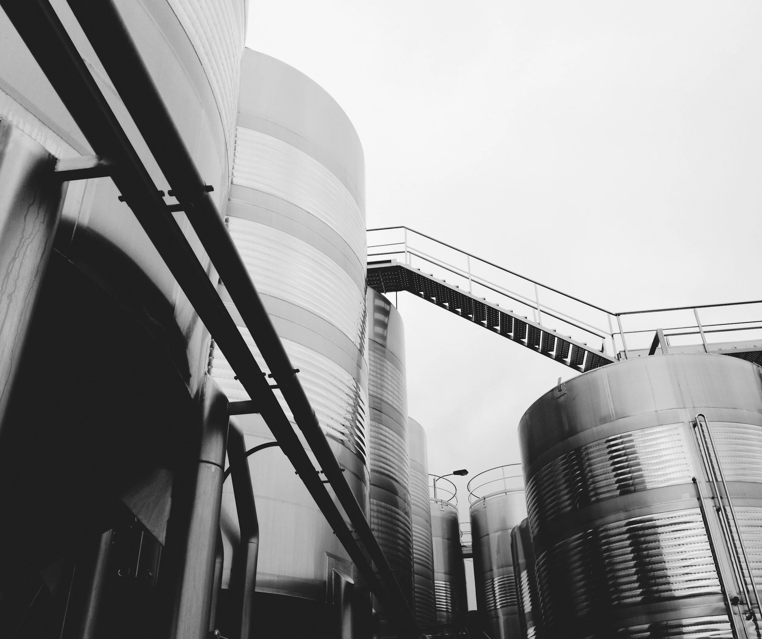 Black and white photo of large stainless steel industrial tanks with a walkway and ladder, likely part of a factory or storage facility.