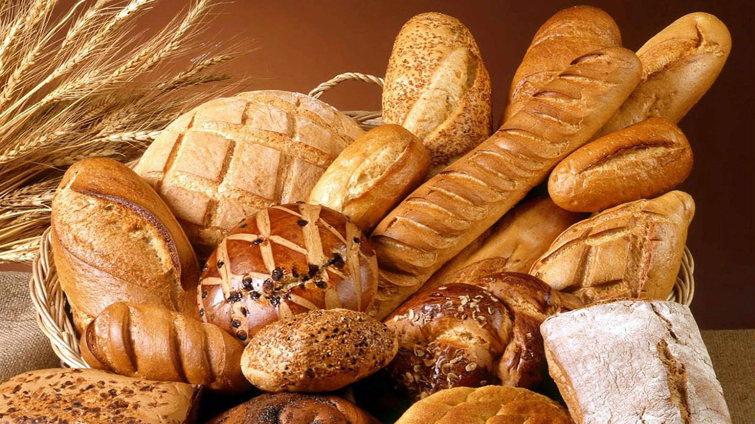 Assorted baked bread loaves and rolls in a basket with wheat stalks in the background.