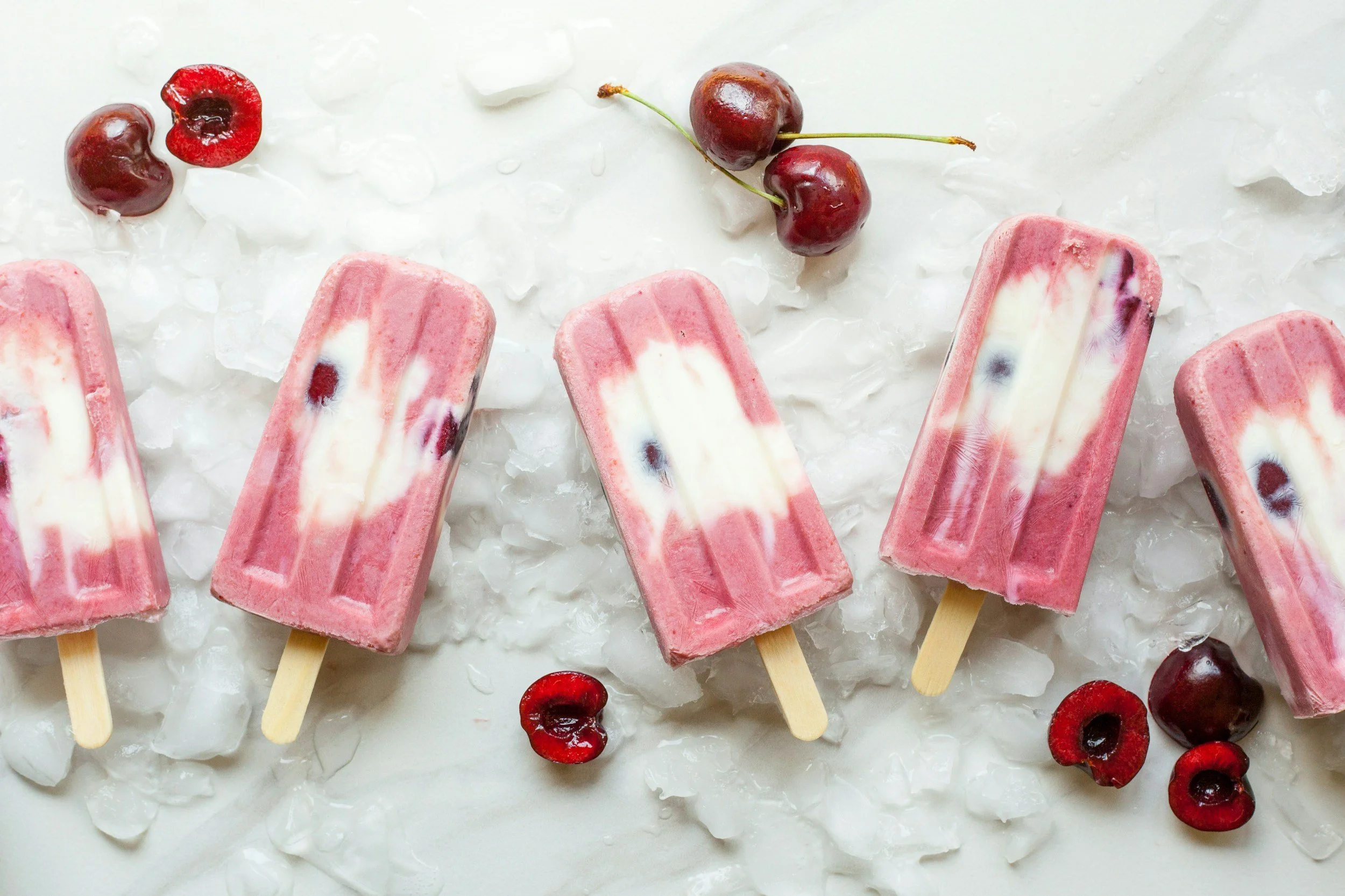 Pink and white ice cream bars on a white surface with ice and cherries scattered around.