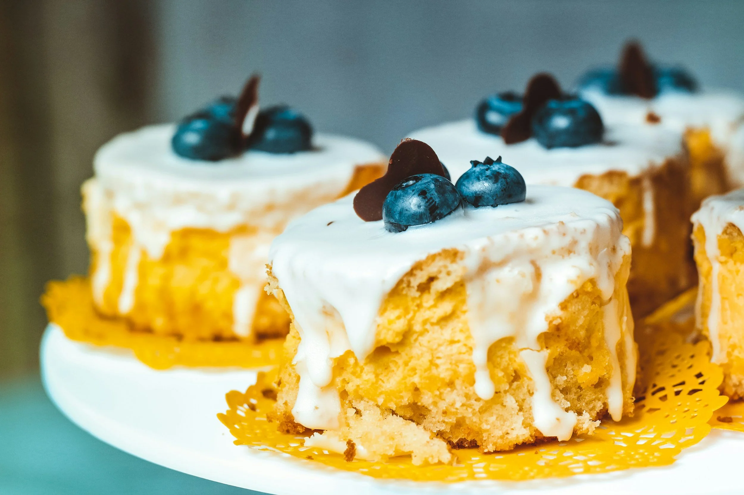 Close-up of slices of blueberry cake with white icing on a cake plate, decorated with fresh blueberries and chocolate pieces.