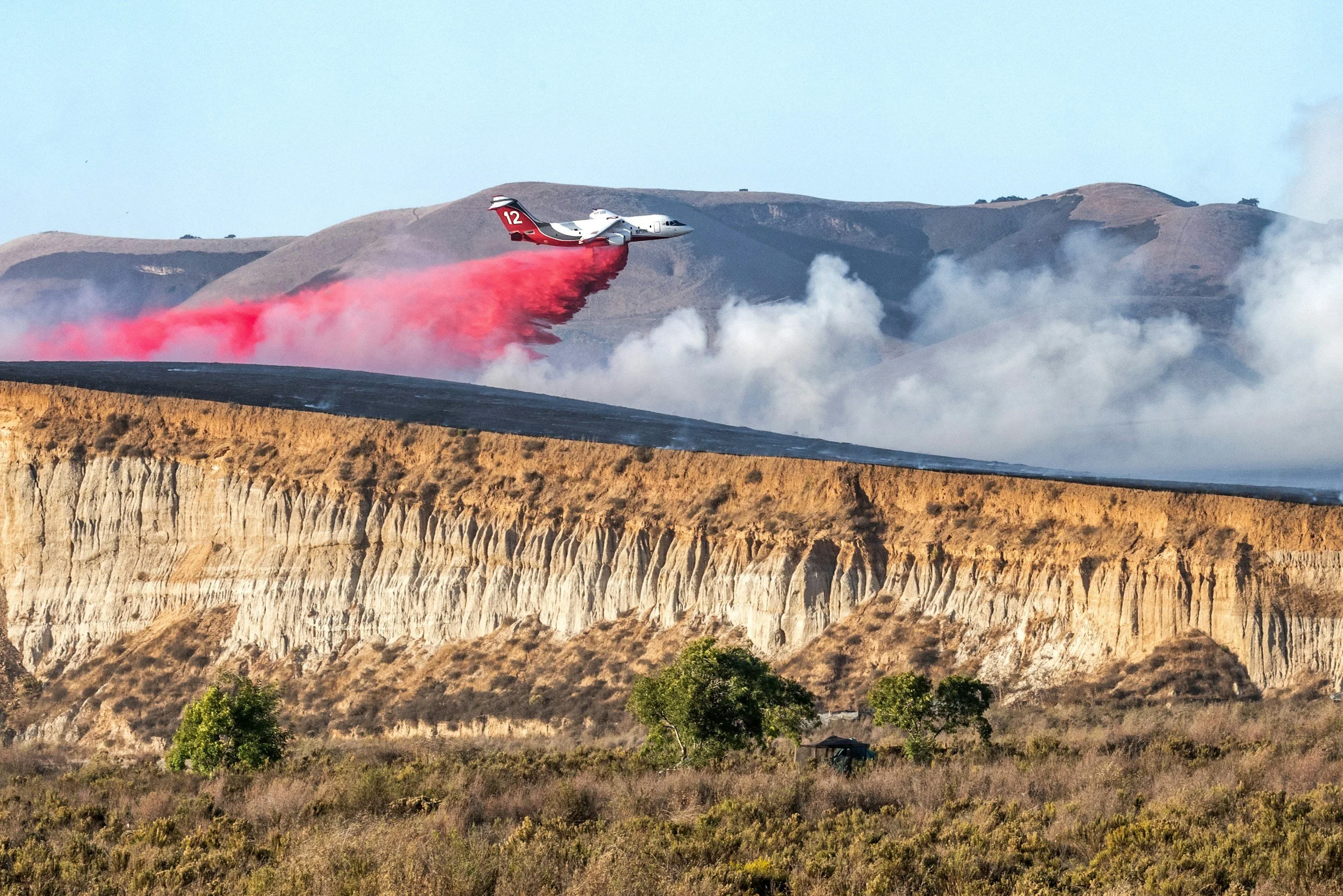 Airplane releasing red fire retardant over a mountainous landscape during a wildfire.