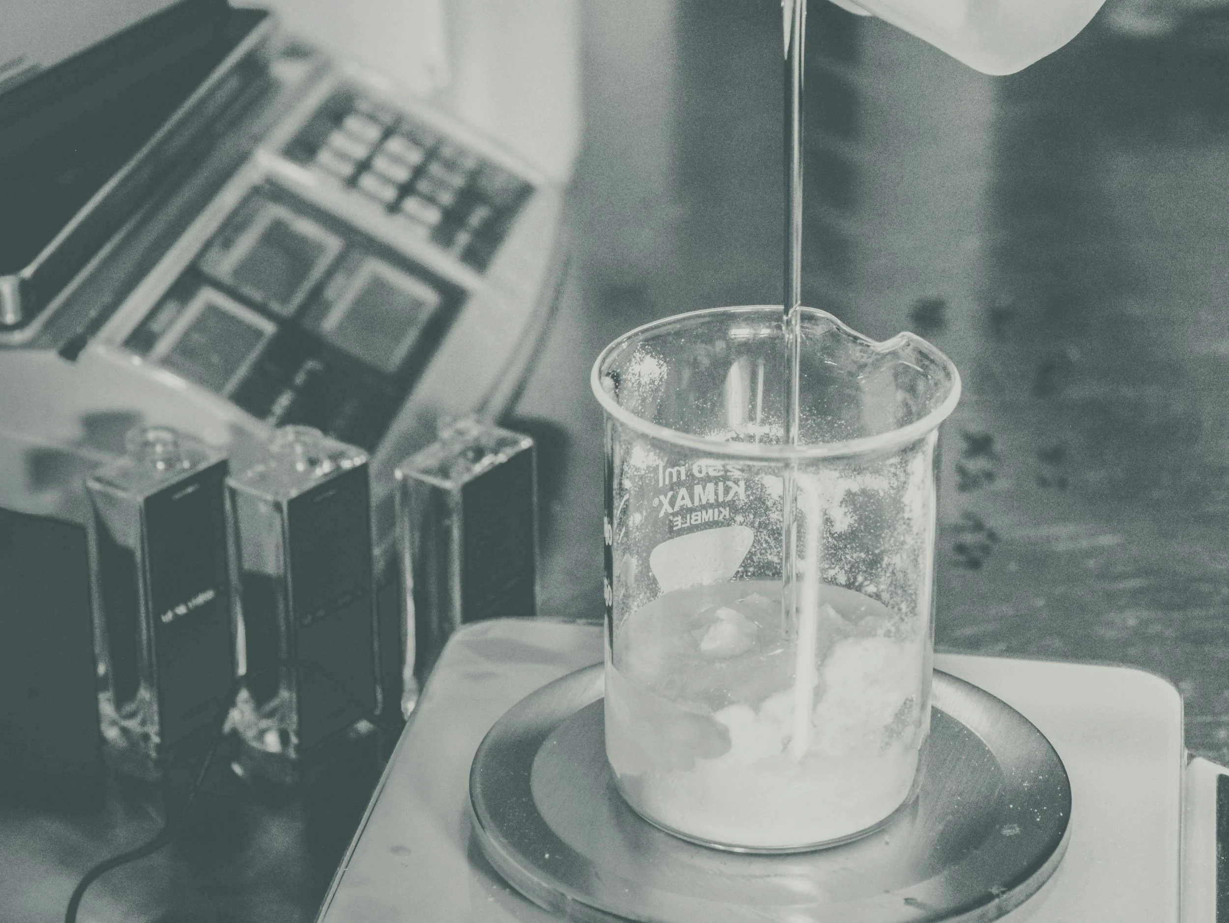 A beaker with a liquid being poured into it, placed on a scale on a work surface.