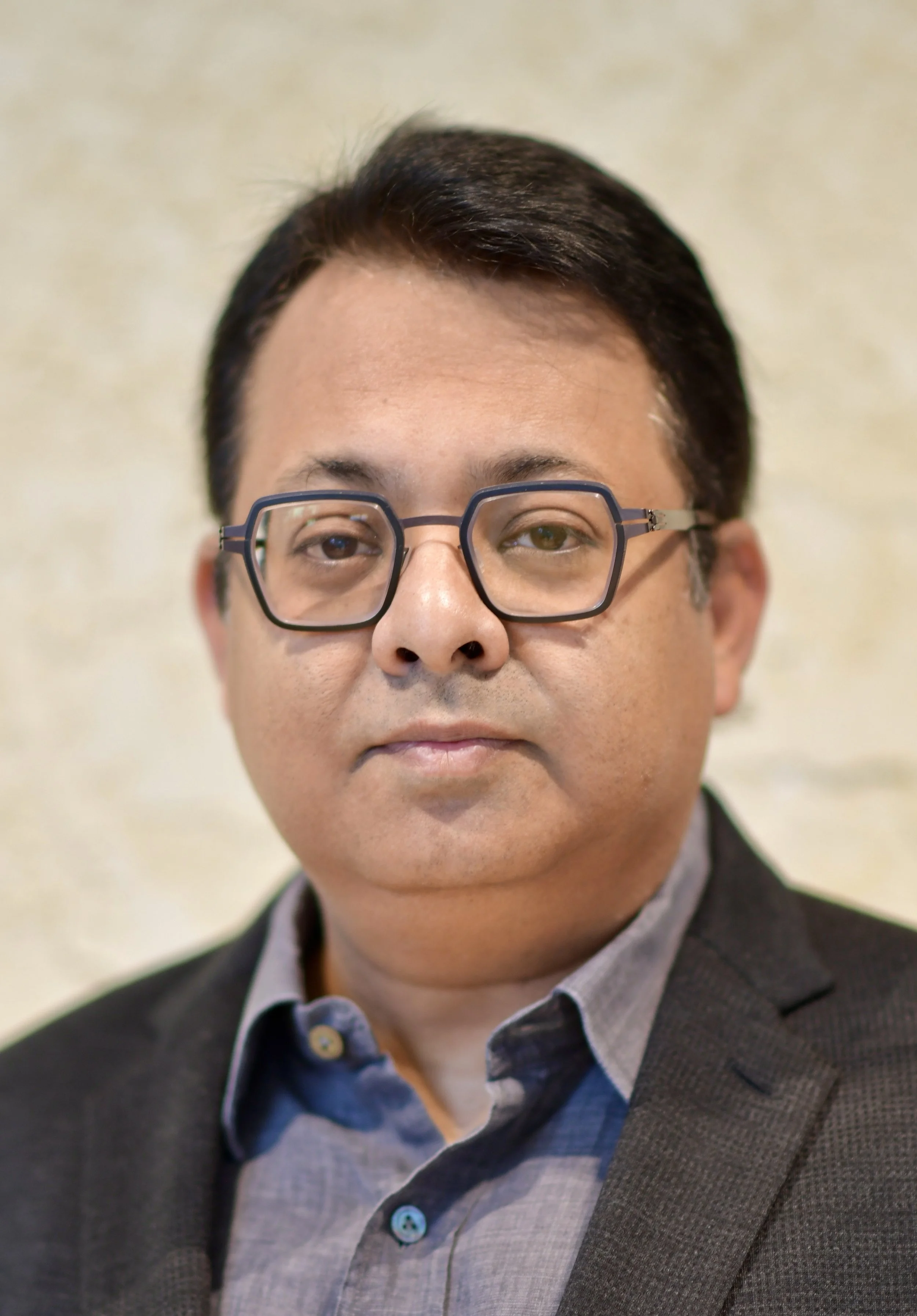 A man with black hair and glasses wearing a suit and collared shirt, posing for a portrait against a light, textured background.