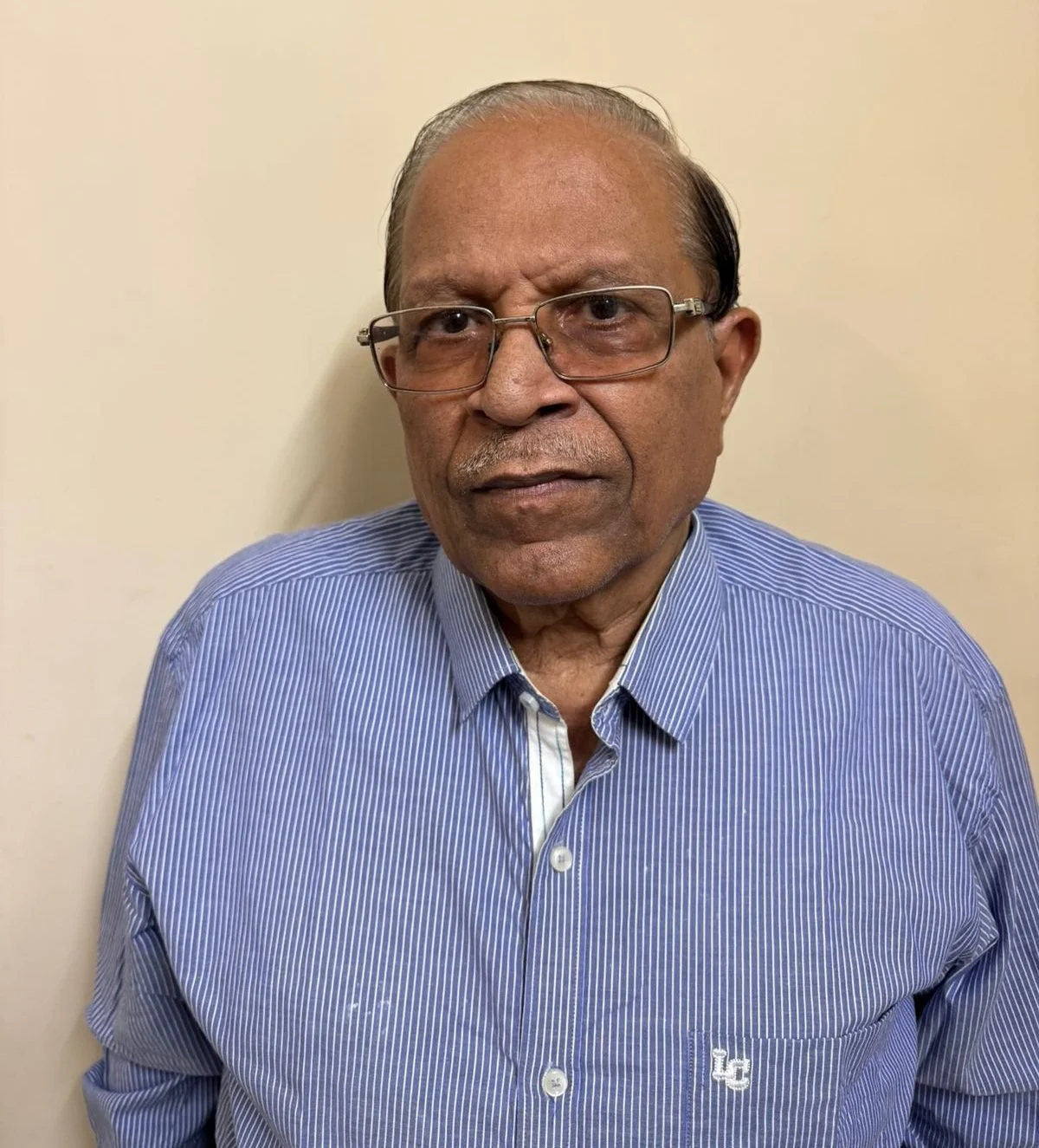 A middle-aged man with glasses and a serious expression wearing a blue and white striped shirt standing against a plain beige wall.