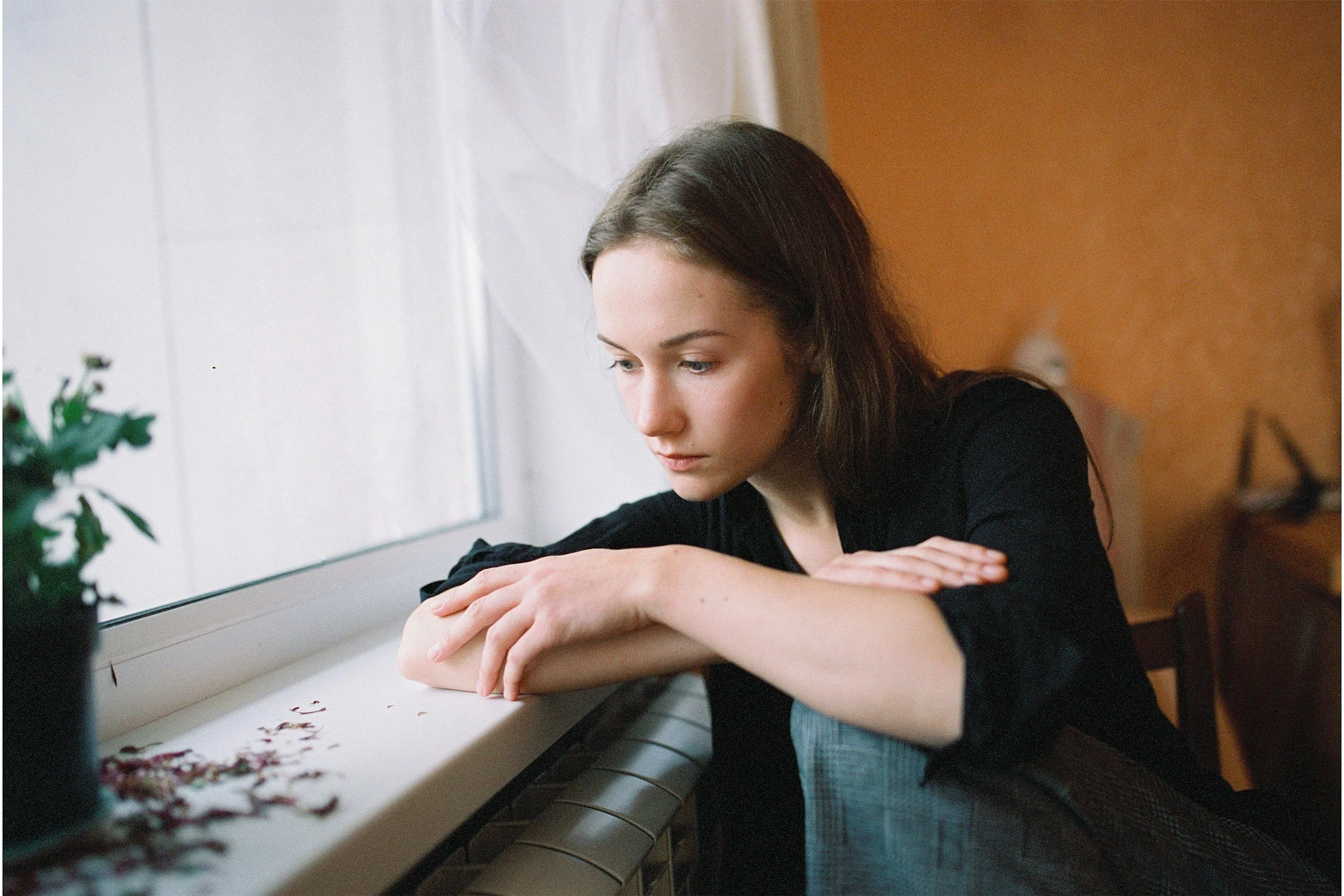 A young woman with brown hair and in a black shirt sits by a window, with her arms resting on the windowsill, looking contemplatively downward. There is a plant on the left side of the image, and the background features a brown wall.