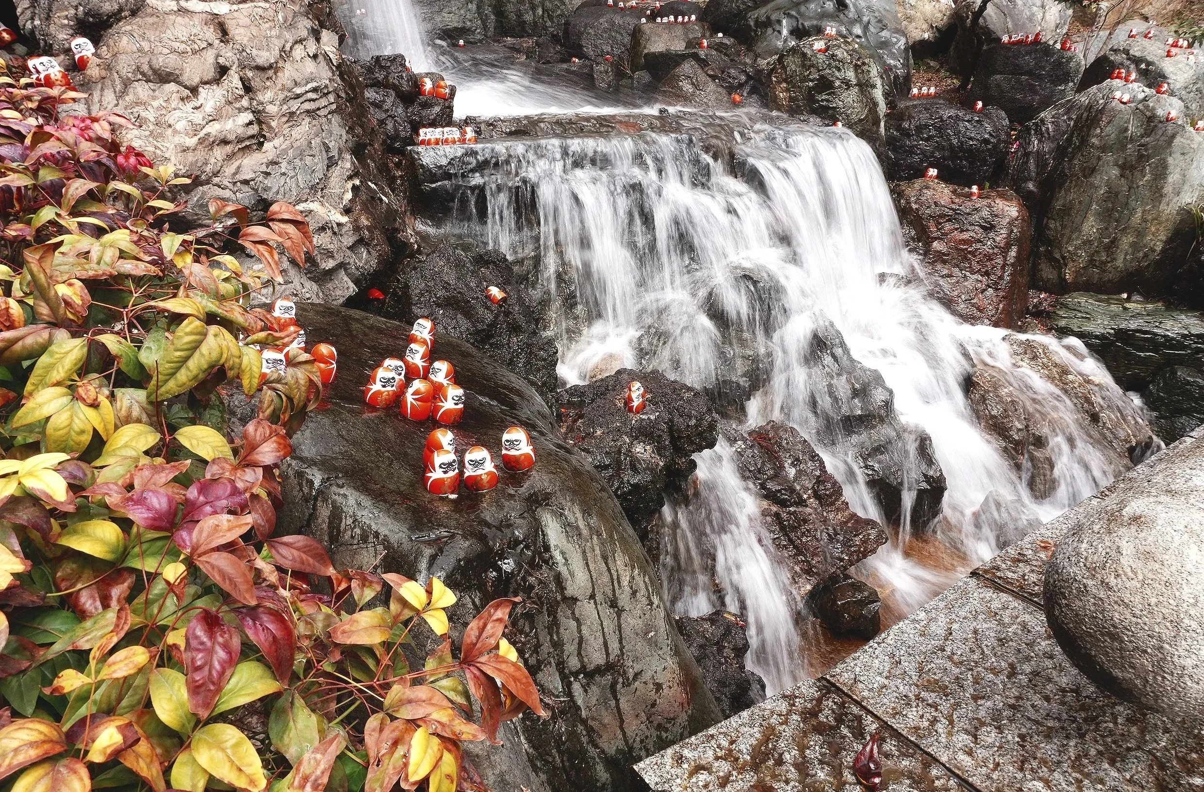 Small decorative Daruma dolls placed on wet rocks around a flowing waterfall in a natural setting with colorful autumn leaves on the left.