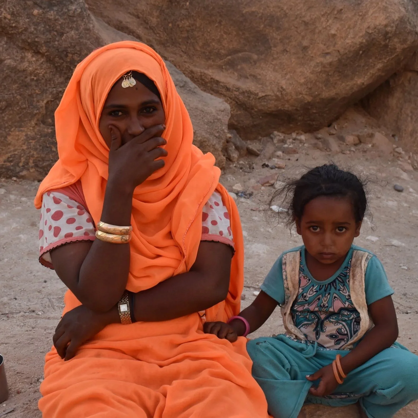 A woman and a girl sitting on the dirt ground in a rocky outdoor setting. The woman is wearing an orange hijab and traditional clothing, and the girl is dressed in a blue outfit with a backpack.