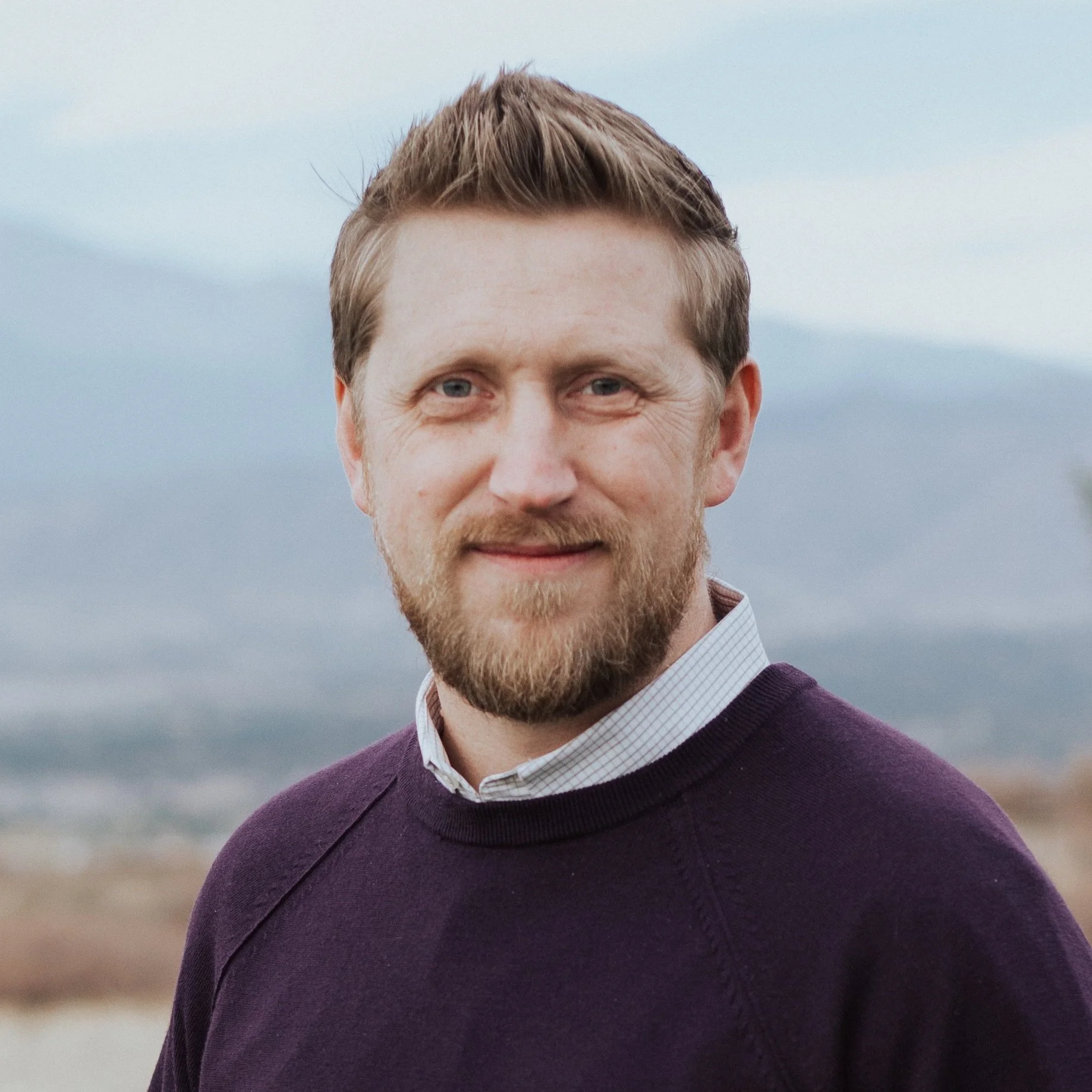 A man with light brown hair, a beard, wearing a purple sweater over a white collared shirt, standing outdoors with mountains and a cloudy sky in the background.