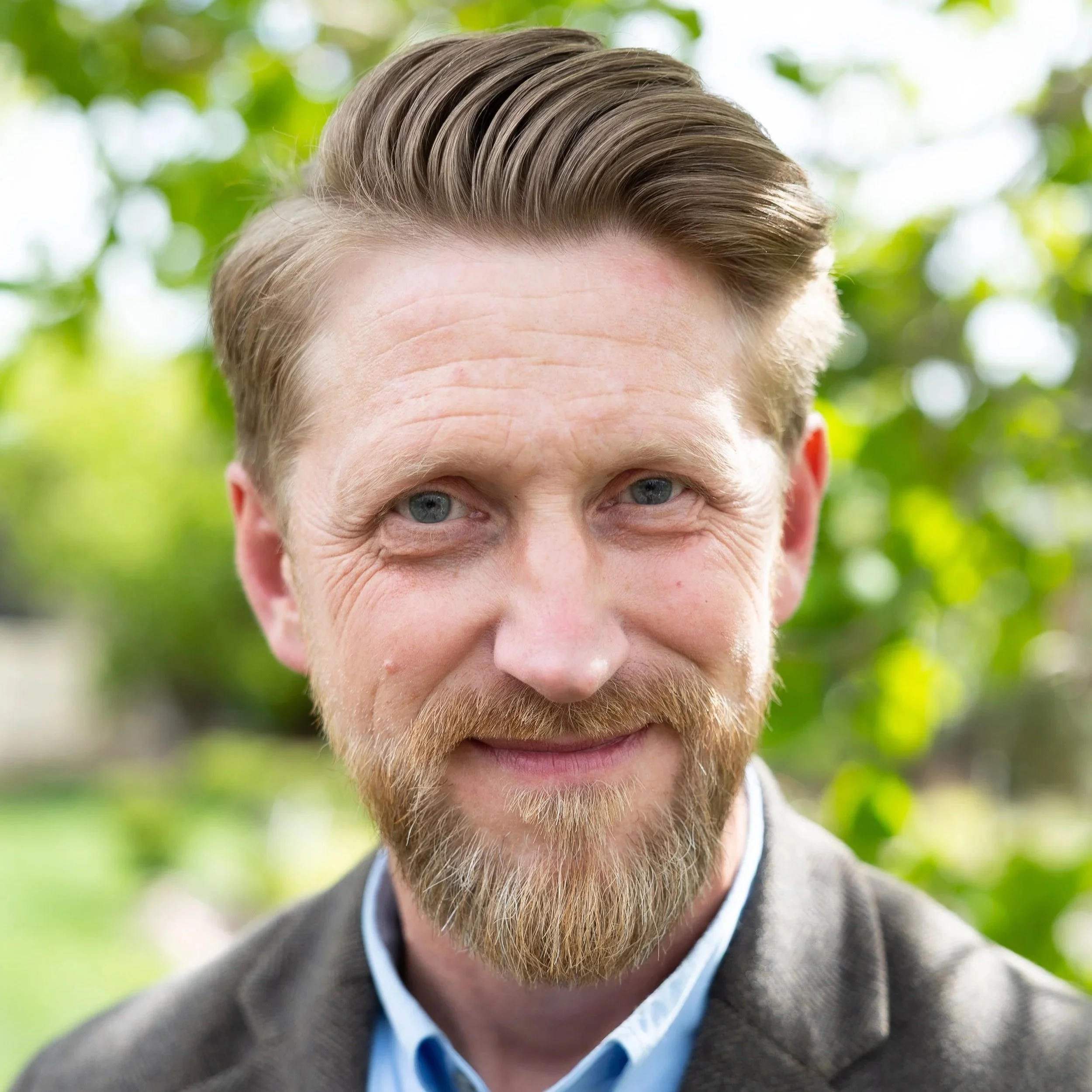 Close-up of a man with blonde hair, blue eyes, and a beard, wearing a gray jacket and light blue shirt, outdoors with green foliage in the background.