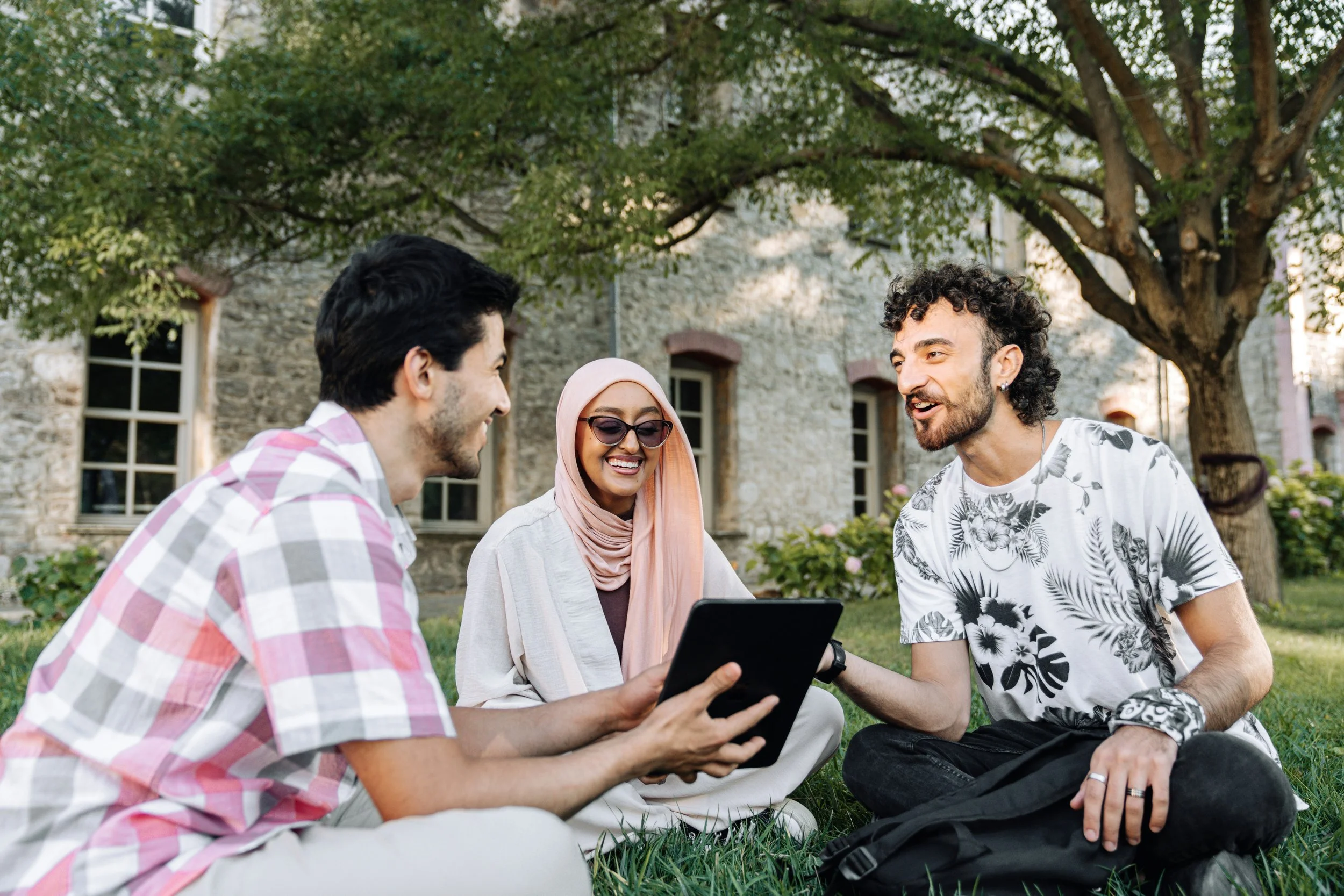 Three young people sitting on grass outside in front of a stone building, smiling and talking, with one holding a tablet.