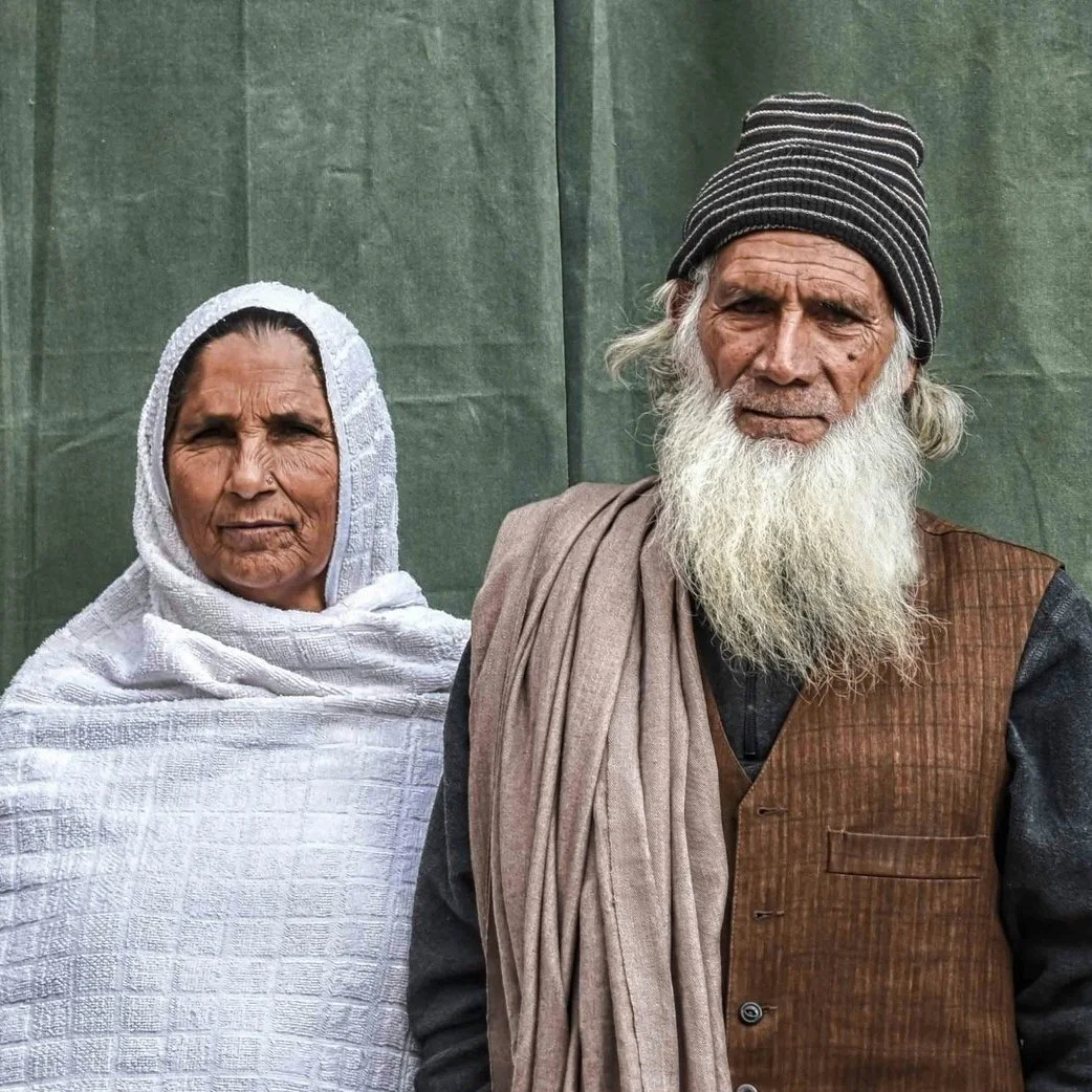 An elderly man with a long white beard and gray hair wearing a beanie, and an elderly woman wearing a white headscarf, standing in front of a green backdrop.