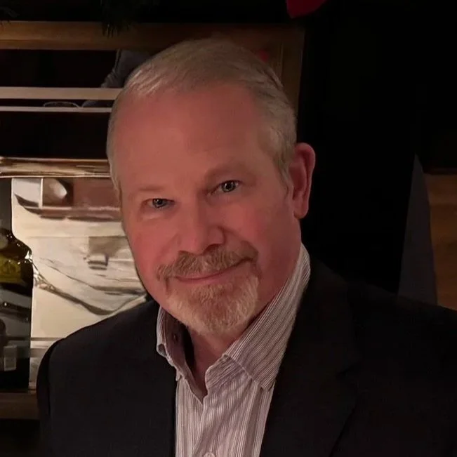 Close-up portrait of a middle-aged man with blonde hair, a beard, and a mustache, smiling, wearing a dark suit and light-colored collared shirt, in an indoor setting.