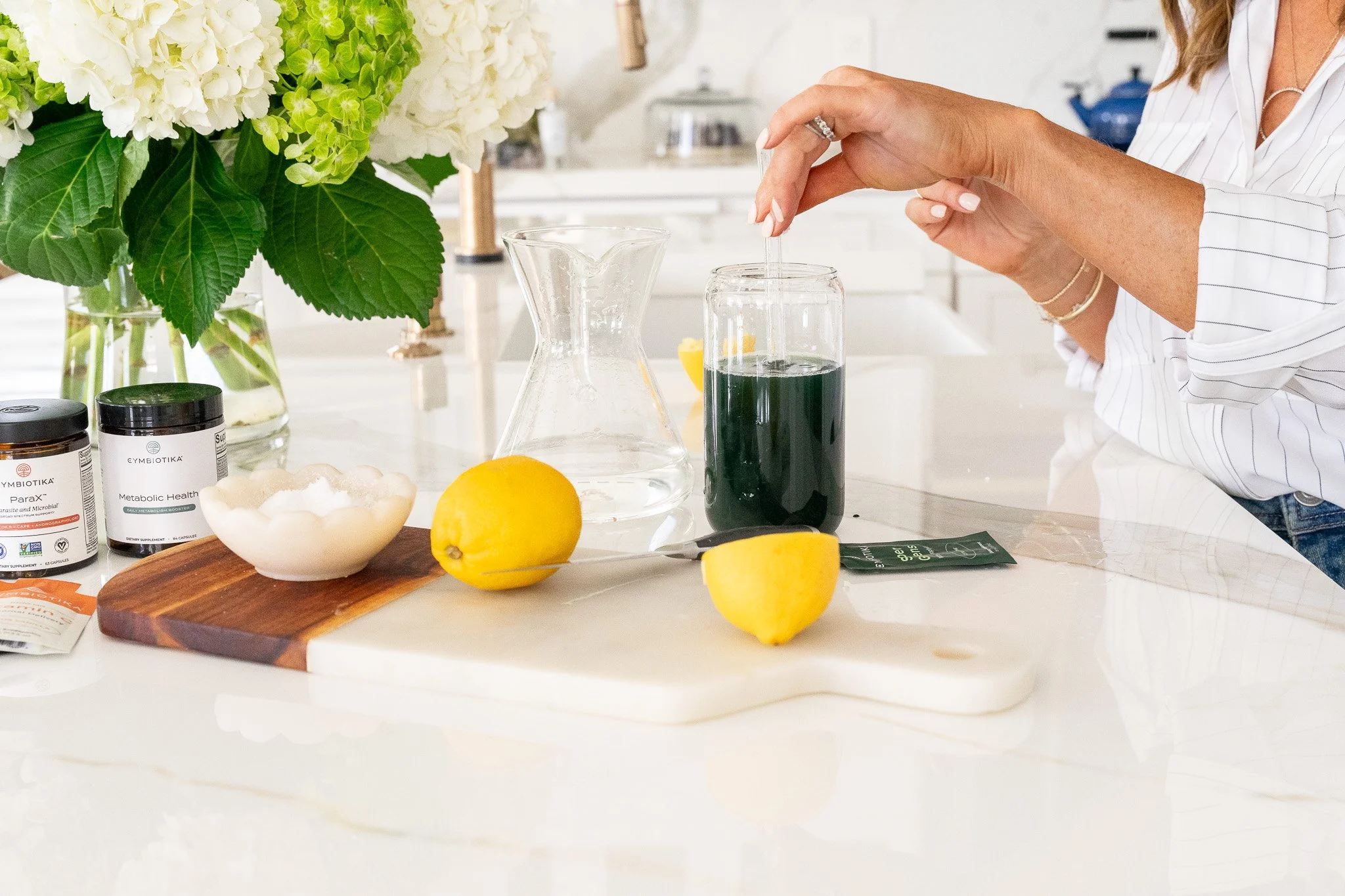 A person stirring a green supplement in a glass jar on a kitchen counter with a marble cutting board, lemons, and nutritional supplements. A vase of white hydrangeas is in the background.