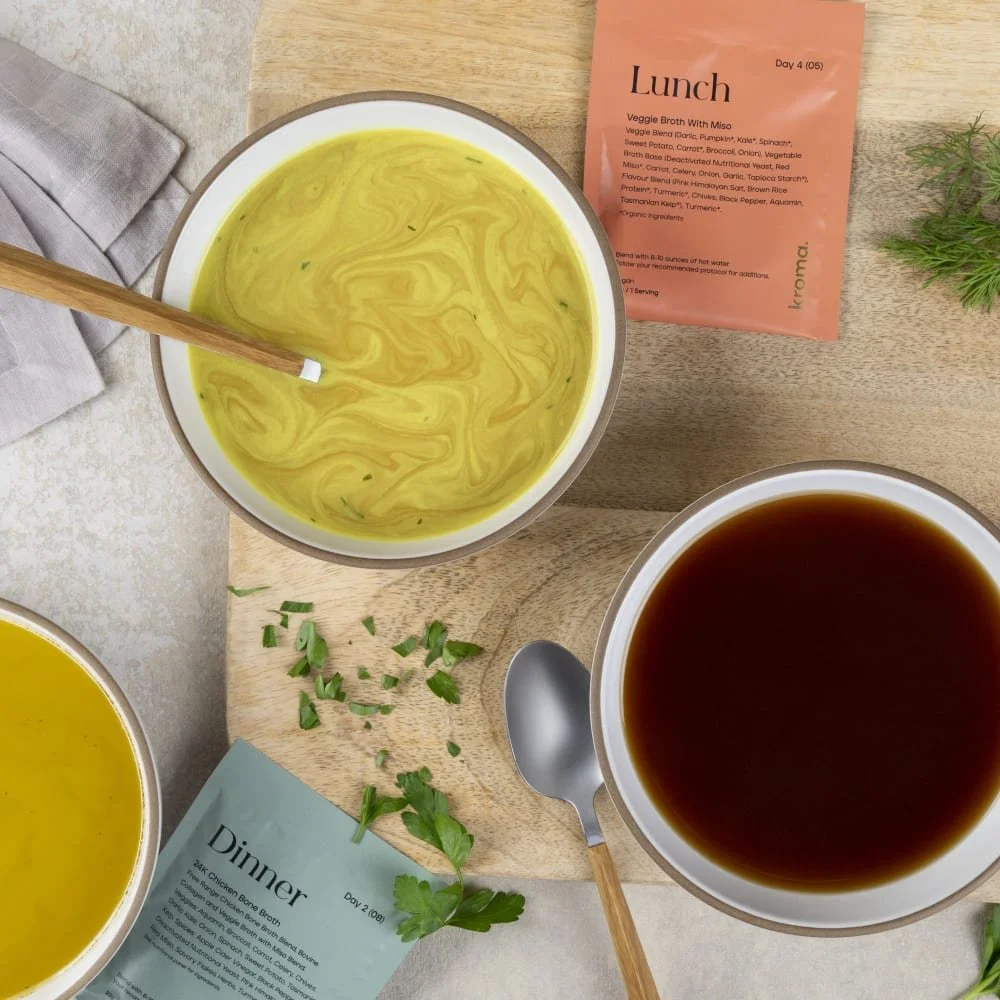 Three bowls of soup on a wooden board with herbs; one is a yellow creamy soup, another is a dark broth, accompanied by labeled lunch and dinner packets from Kroma 5 Day Reset.