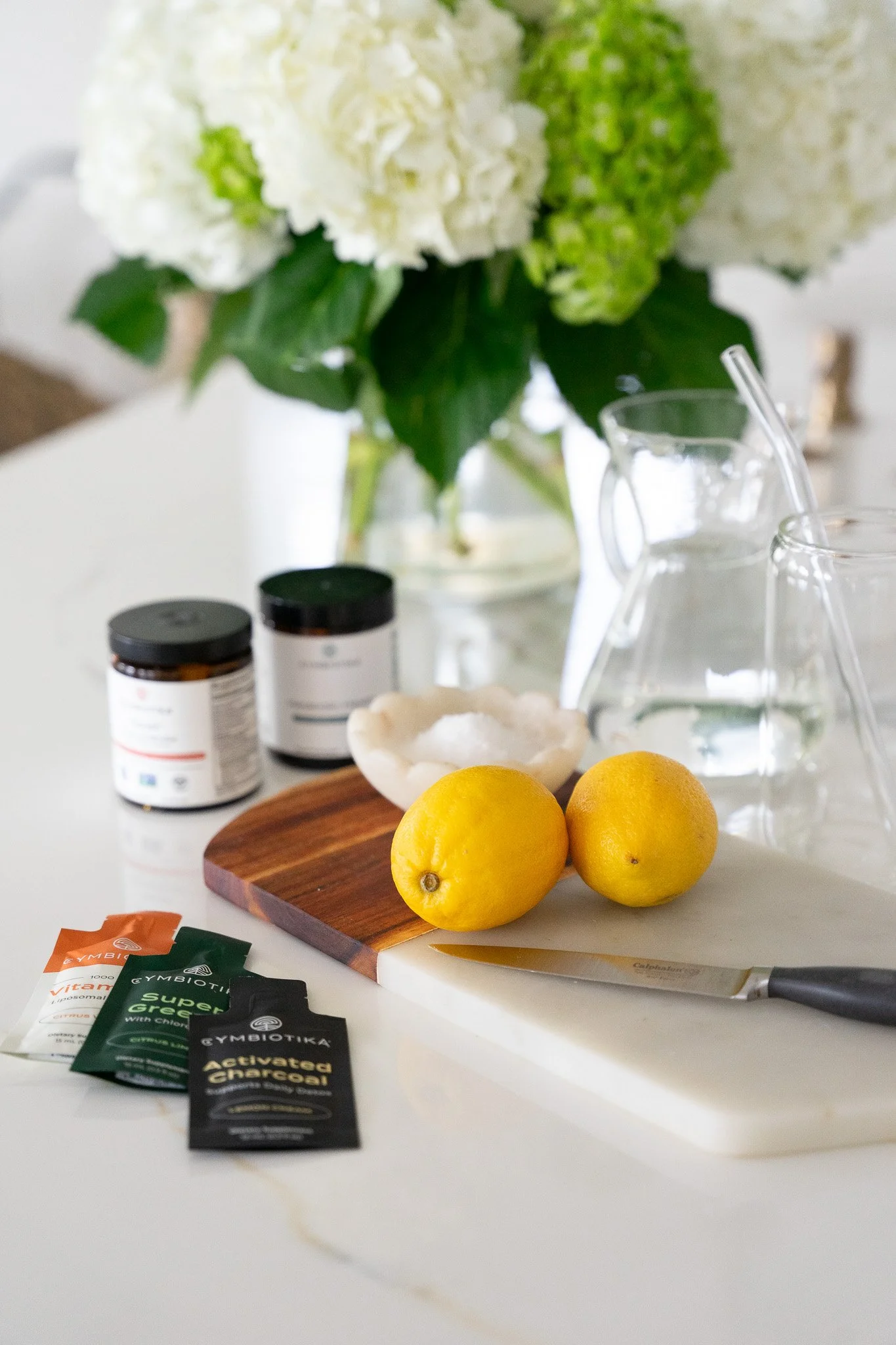 White countertop with lemons on cutting board, knife, water in glass pitchers, supplement packets, jars, and decorative flowers.
