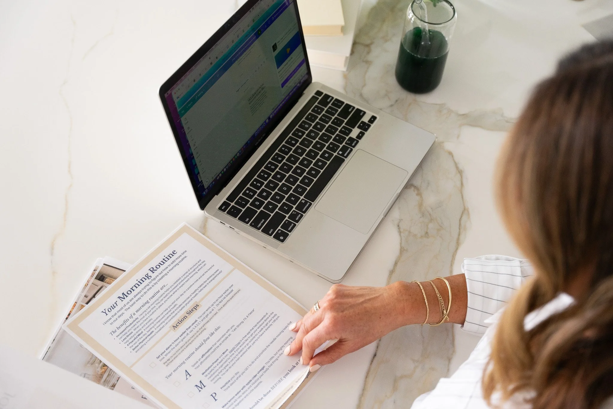 Overhead view of a person sitting at a desk working on a laptop with documents titled "Your Morning Routine" next to a green drink in a cup.