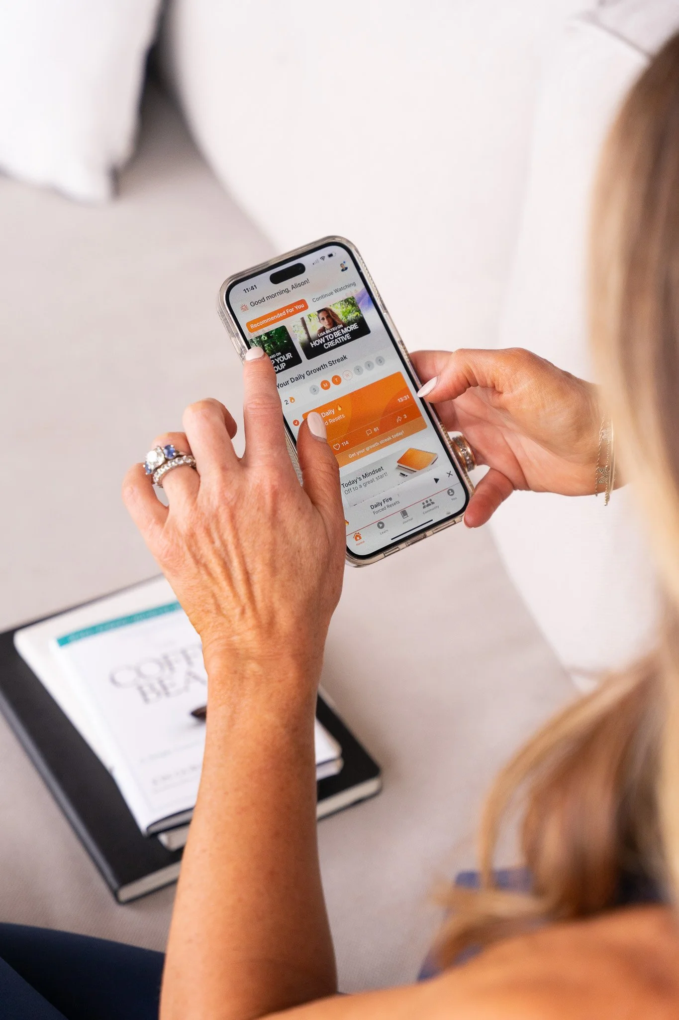 Person using a smartphone app, with a book titled "The Coffee Bean" nearby on a couch.