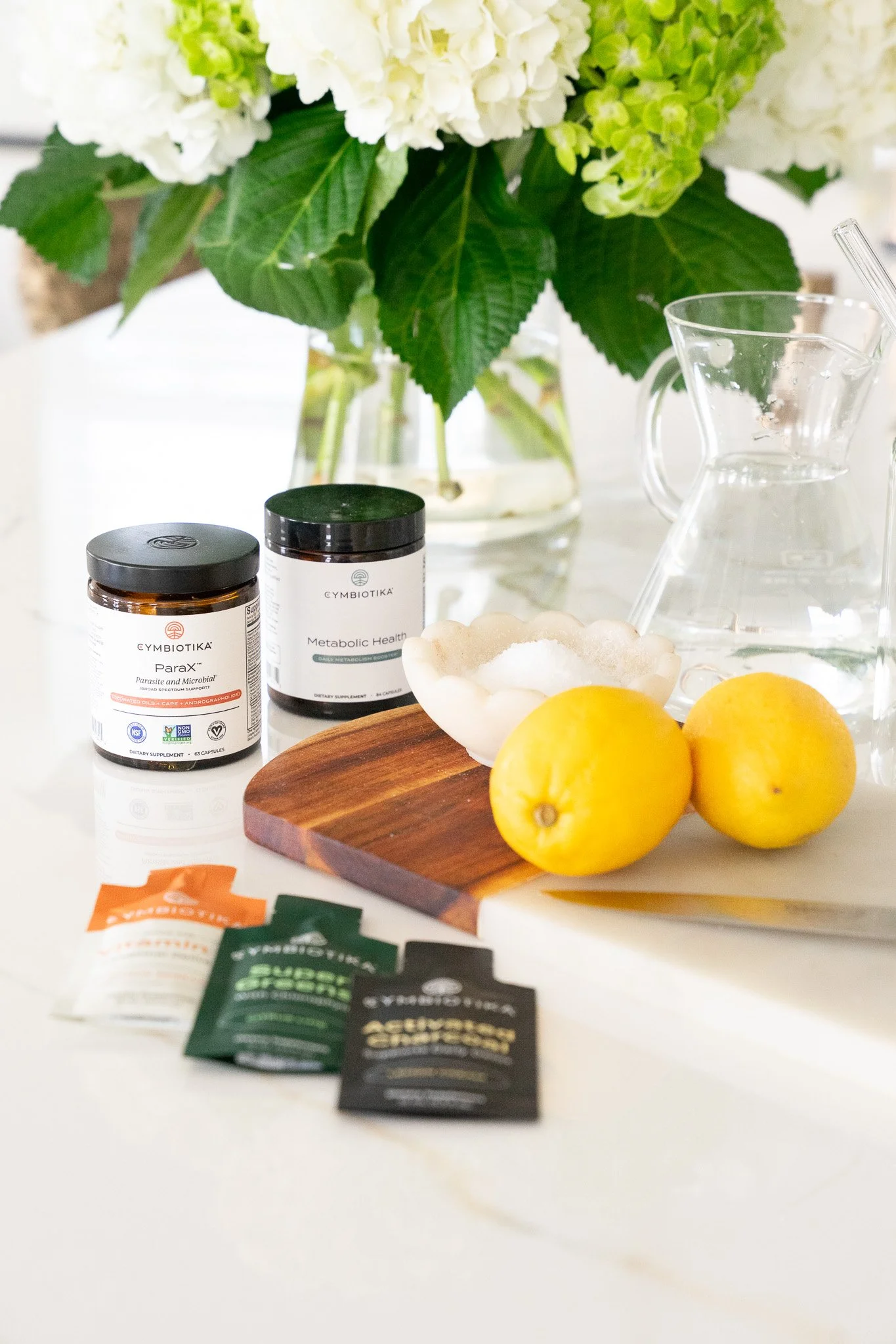 Health supplements and ingredients on a kitchen counter from Cymbiotica, a bowl with white powder, lemons, supplement sachets, and a pitcher, with hydrangea flowers in the background.
