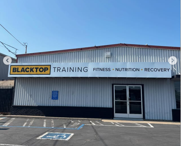 Gray metal building with a banner reading 'BLACKTOP TRAINING FITNESS NUTRITION RECOVERY' and marked handicap parking spaces in front.
