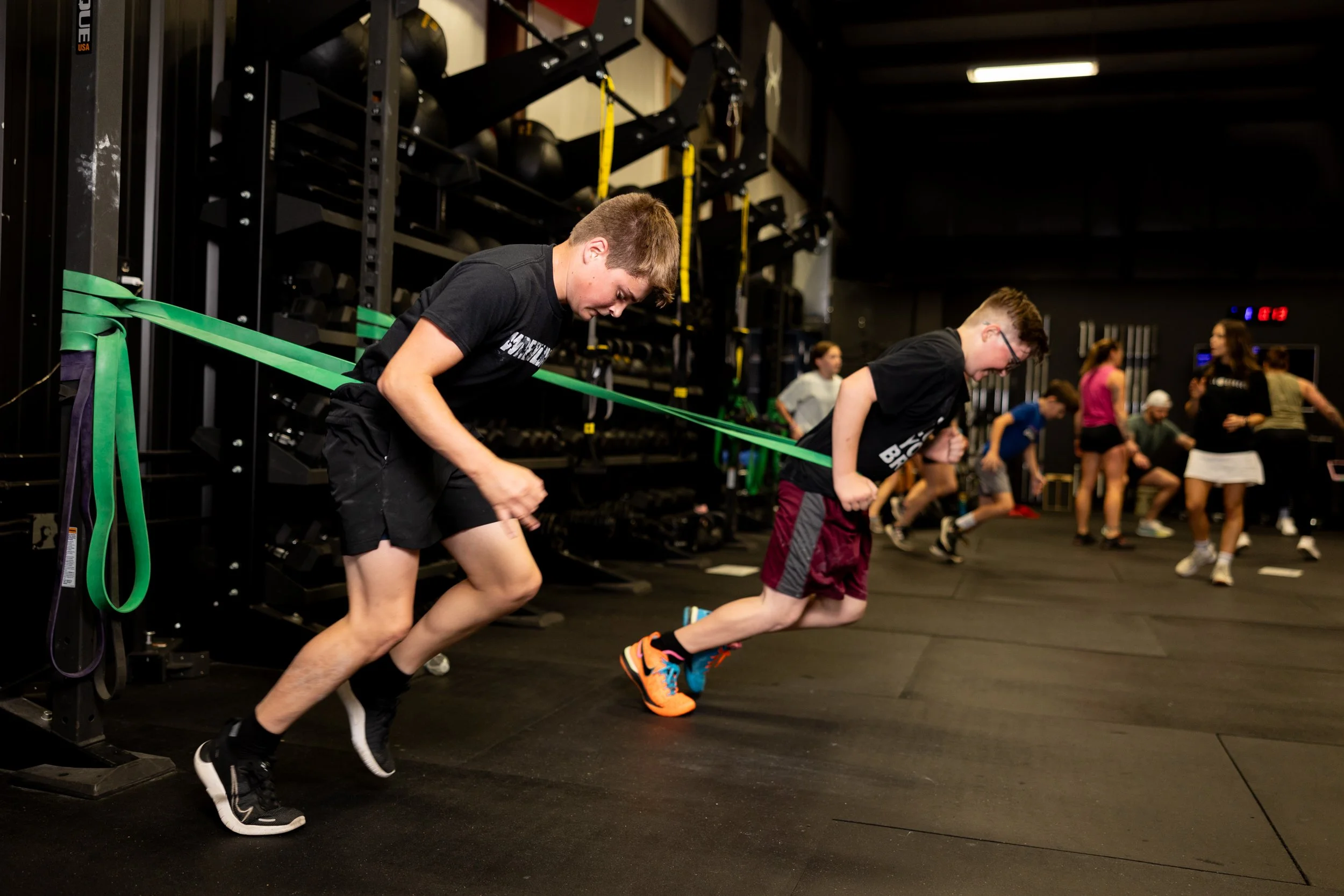 Two young men in black T-shirts and shorts performing resistance band exercises in a gym. Several other people are working out in the background.