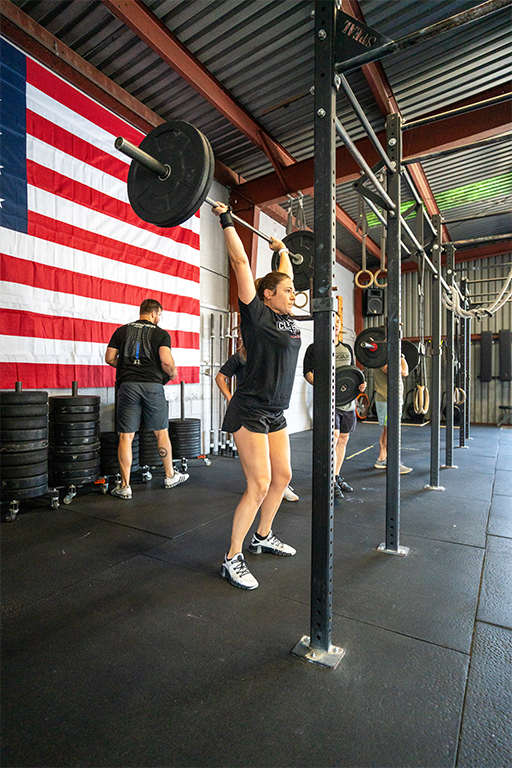 Woman lifting a barbell overhead during a CrossFit workout in a gym with an American flag on the wall.