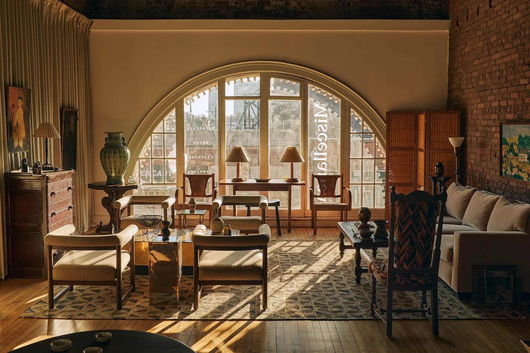 Living room with large arched window, sunlight, vintage furniture, paintings, and brick wall.