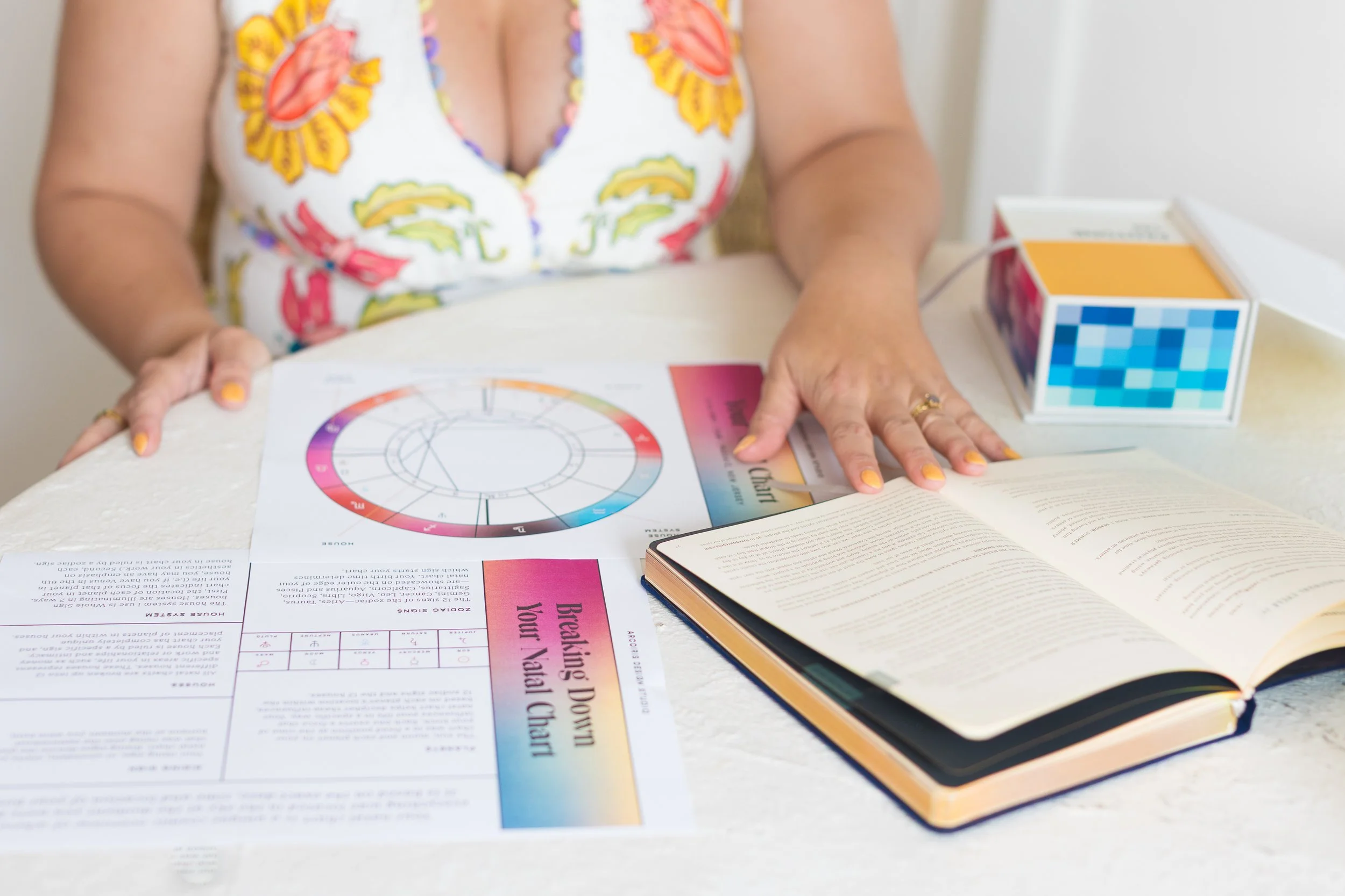 A woman in a colorful dress sitting at a table, using an astrology wheel, an open book, and a box of colorful blocks.
