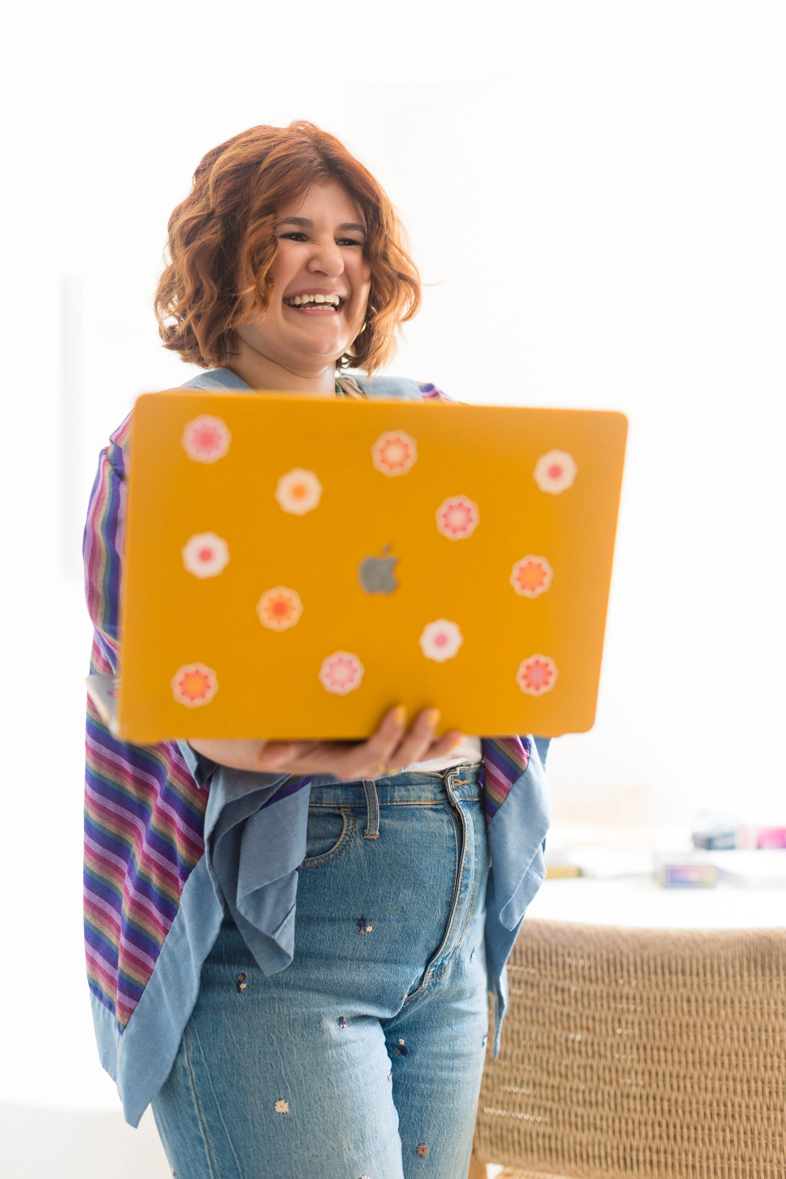 A woman with curly red hair holding a yellow laptop with pink flower stickers, smiling and standing indoors.
