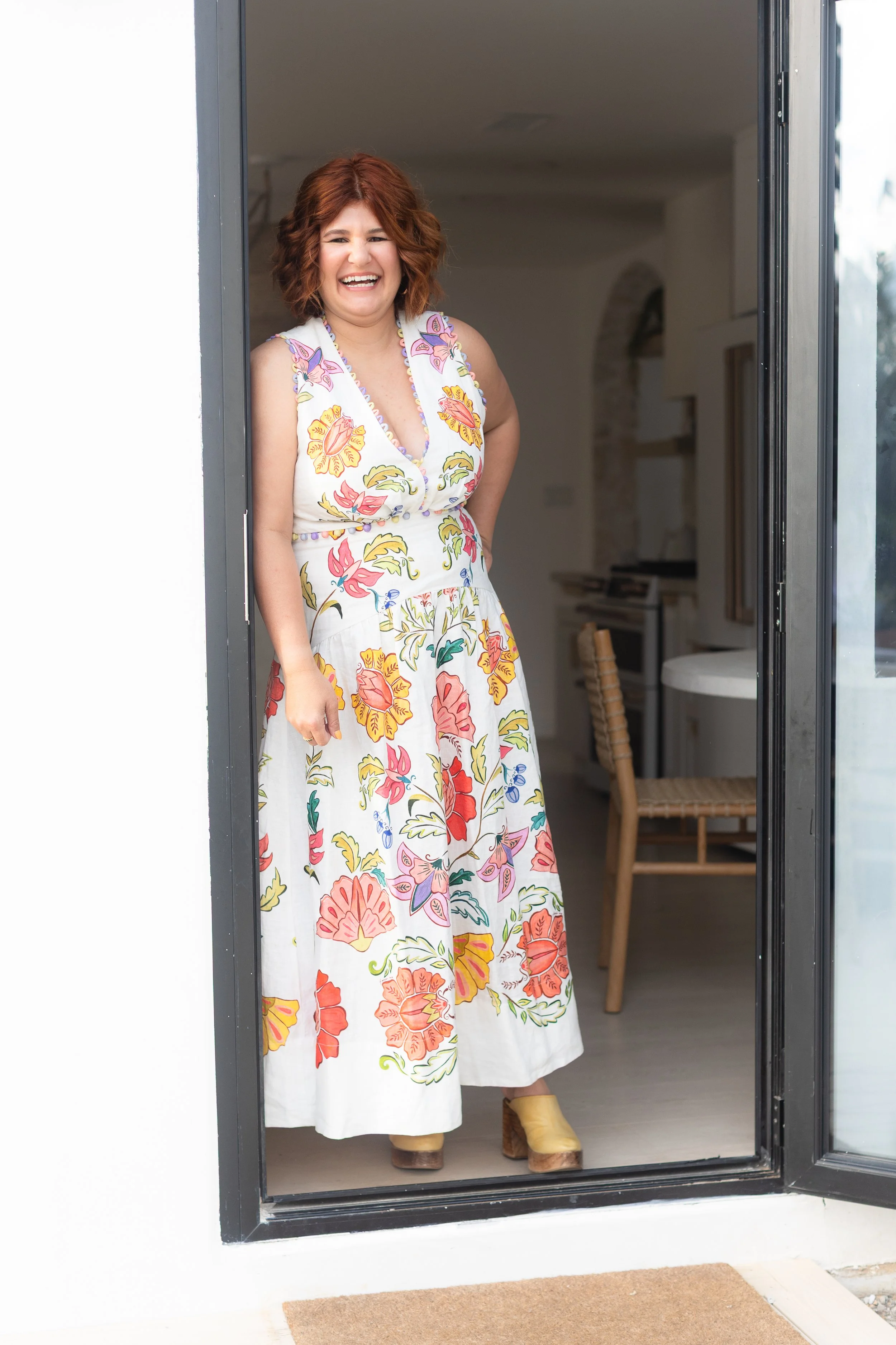 A woman with curly red hair, wearing a colorful floral dress and tan platform shoes, standing at a doorway and smiling.