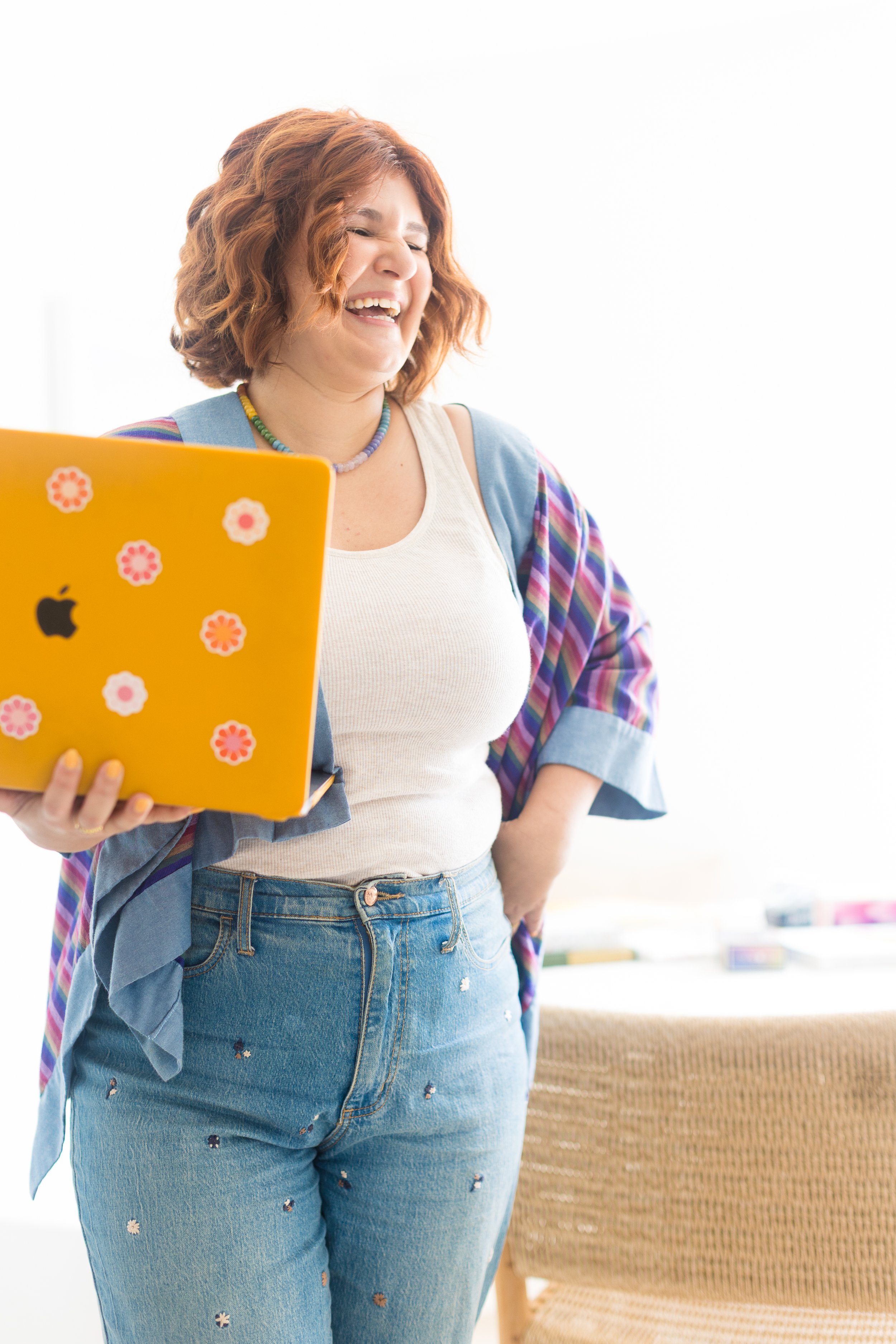 A woman with curly red hair smiling and laughing while holding a yellow laptop decorated with stickers.