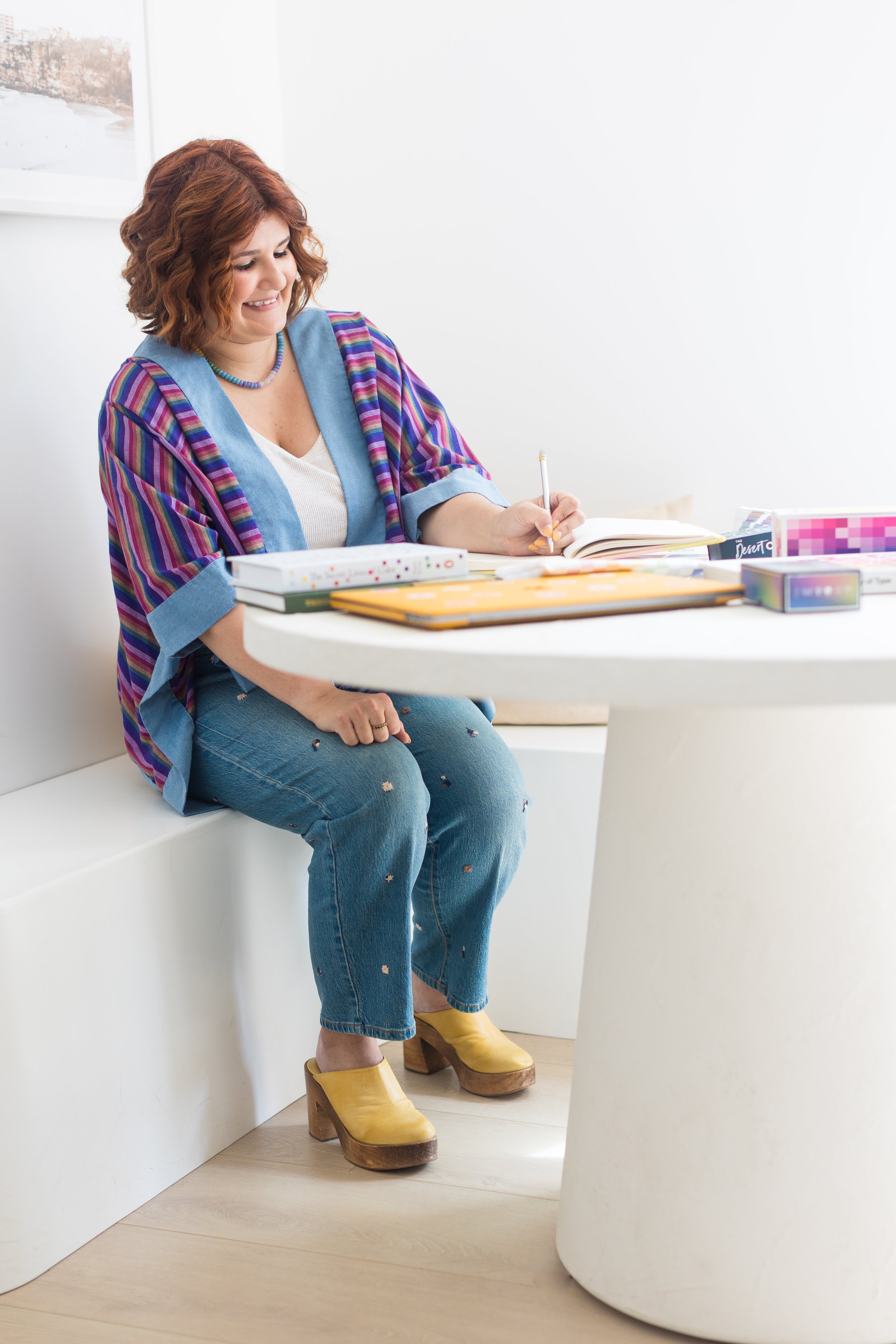 A woman with red hair wearing a multicolored striped jacket, blue jeans with small embellishments, and yellow platform shoes, sitting at a white rounded table, smiling, writing in a notebook, with colorful stationery items on the table.