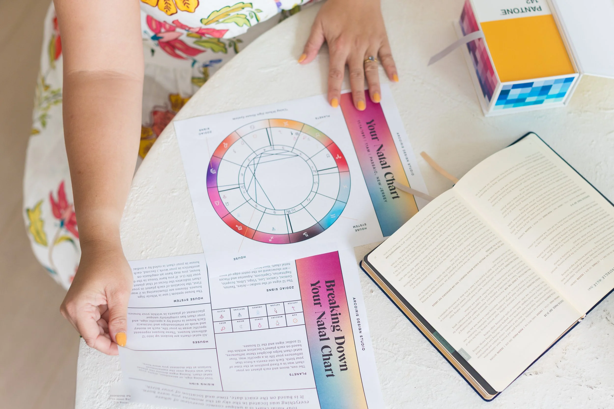 A person with yellow-painted nails working on a colorful natal chart called 'Your Valal Chart', on a white textured table. There is an open book and a multicolored cube on the table.