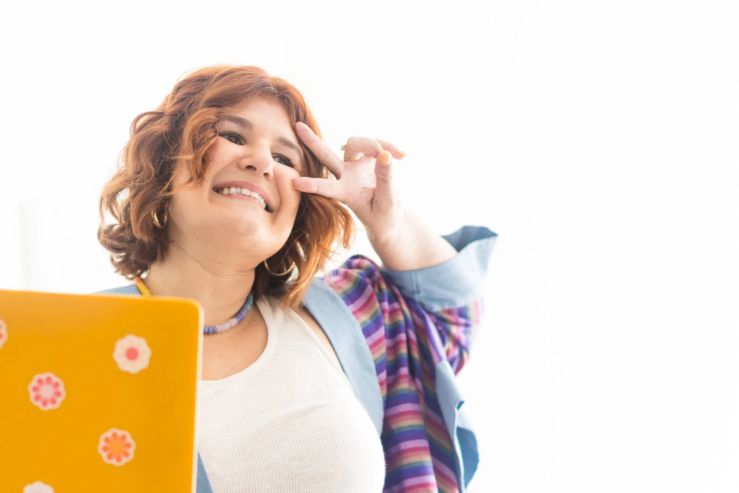 A young woman with curly red hair smiling and making a peace sign near her face, holding a yellow folder with flower designs, against a bright white background.