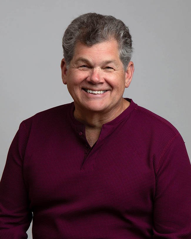 A smiling middle-aged man wearing a maroon shirt, standing against a plain gray background.