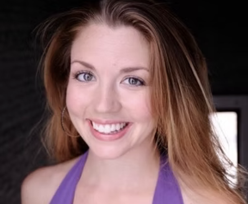 A young woman with red hair, blue eyes, and a bright smile, wearing a purple top.