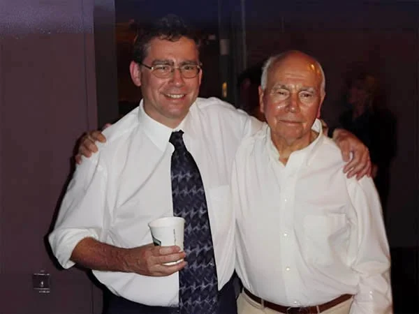 Two men with arms around each other, smiling. The man on the left is holding a white cup, wearing glasses, a white shirt, and a tie. The man on the right is older, with white hair, wearing a white shirt and beige pants.