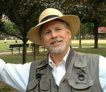 A man with a gray beard wearing a wide-brimmed hat and a light-colored vest, standing outdoors in a park with trees and a bench in the background.